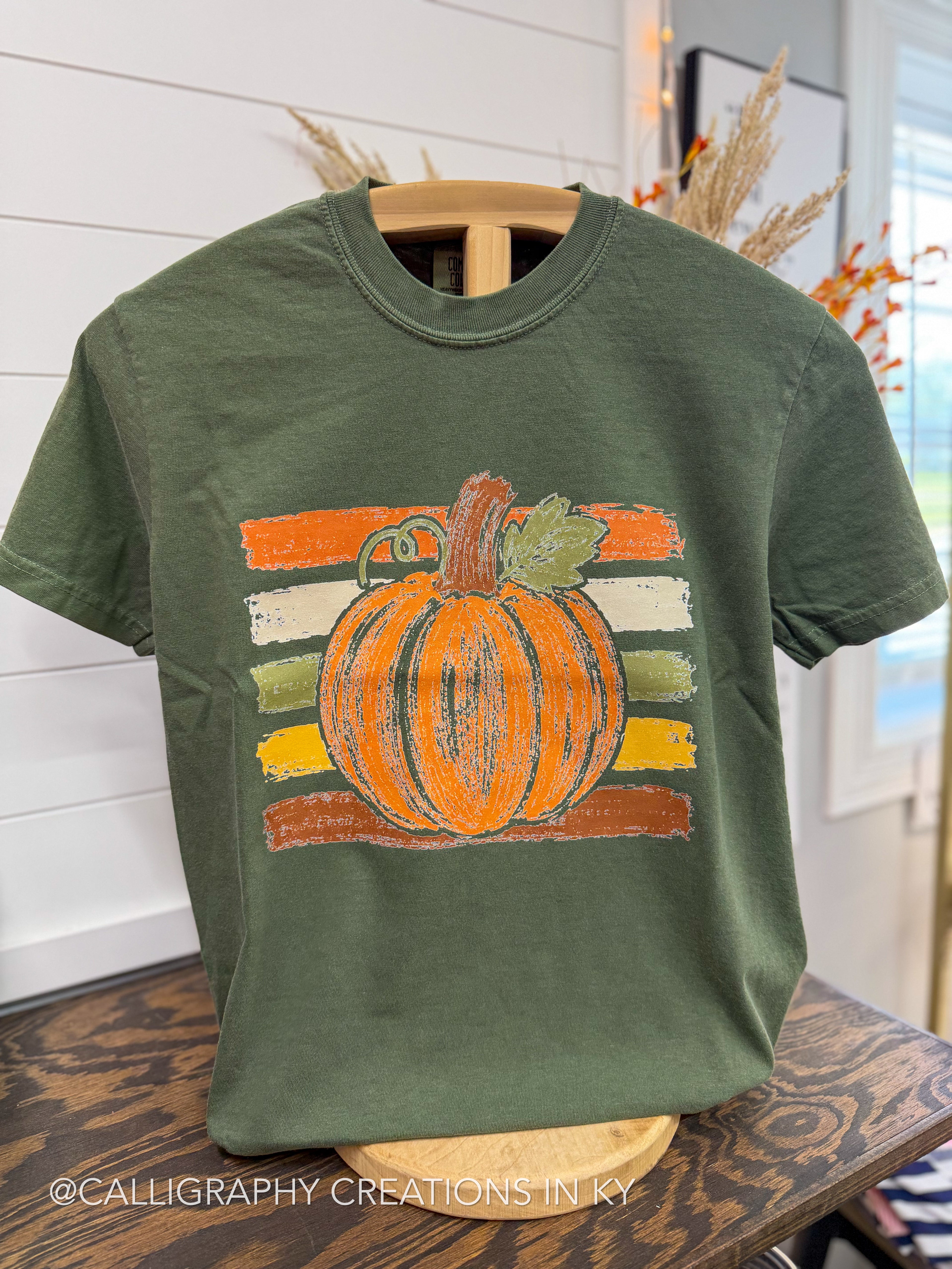 An olive green short-sleeve t-shirt on a wooden hanger, featuring a distressed orange pumpkin graphic with a green leaf and striped background details.