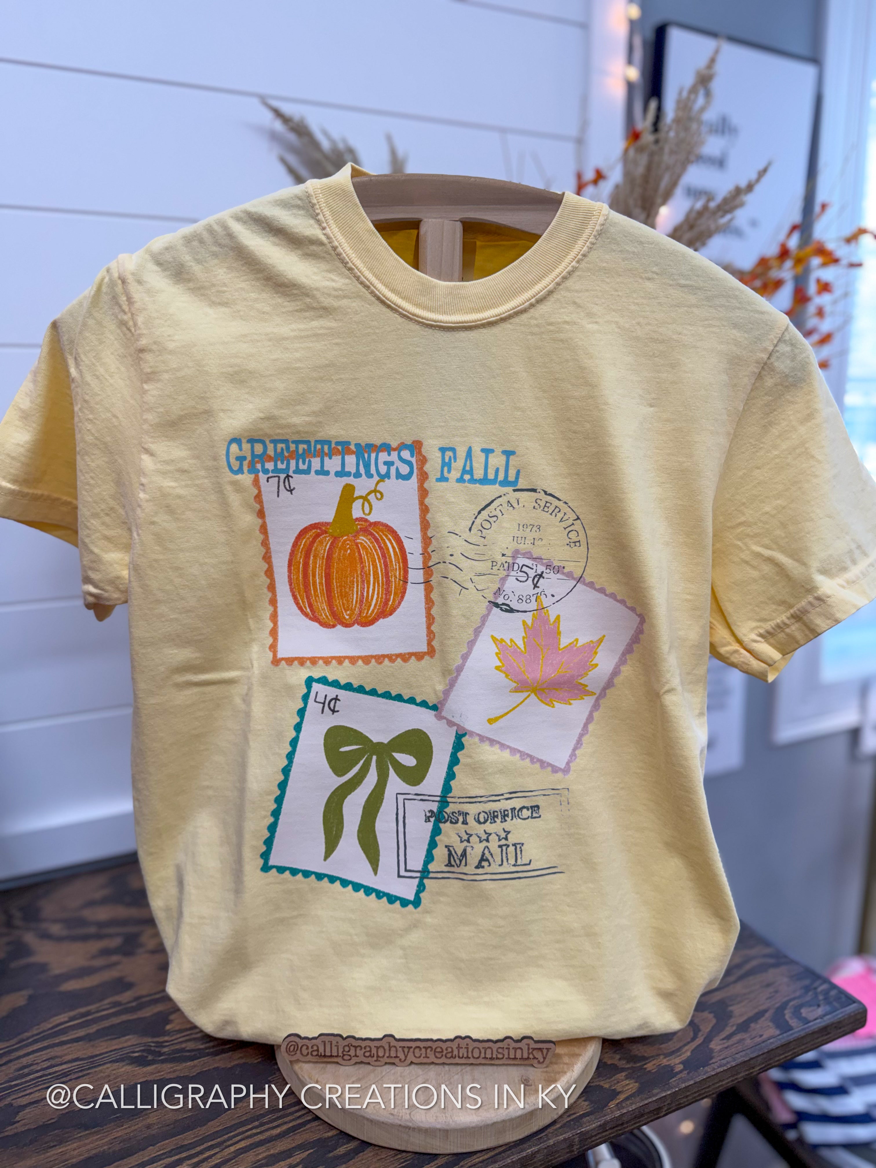 A pale yellow short-sleeve t-shirt with a 'Greetings Fall' graphic print featuring autumn-themed postage stamps, a pumpkin, and a maple leaf, displayed on a wooden hanger.