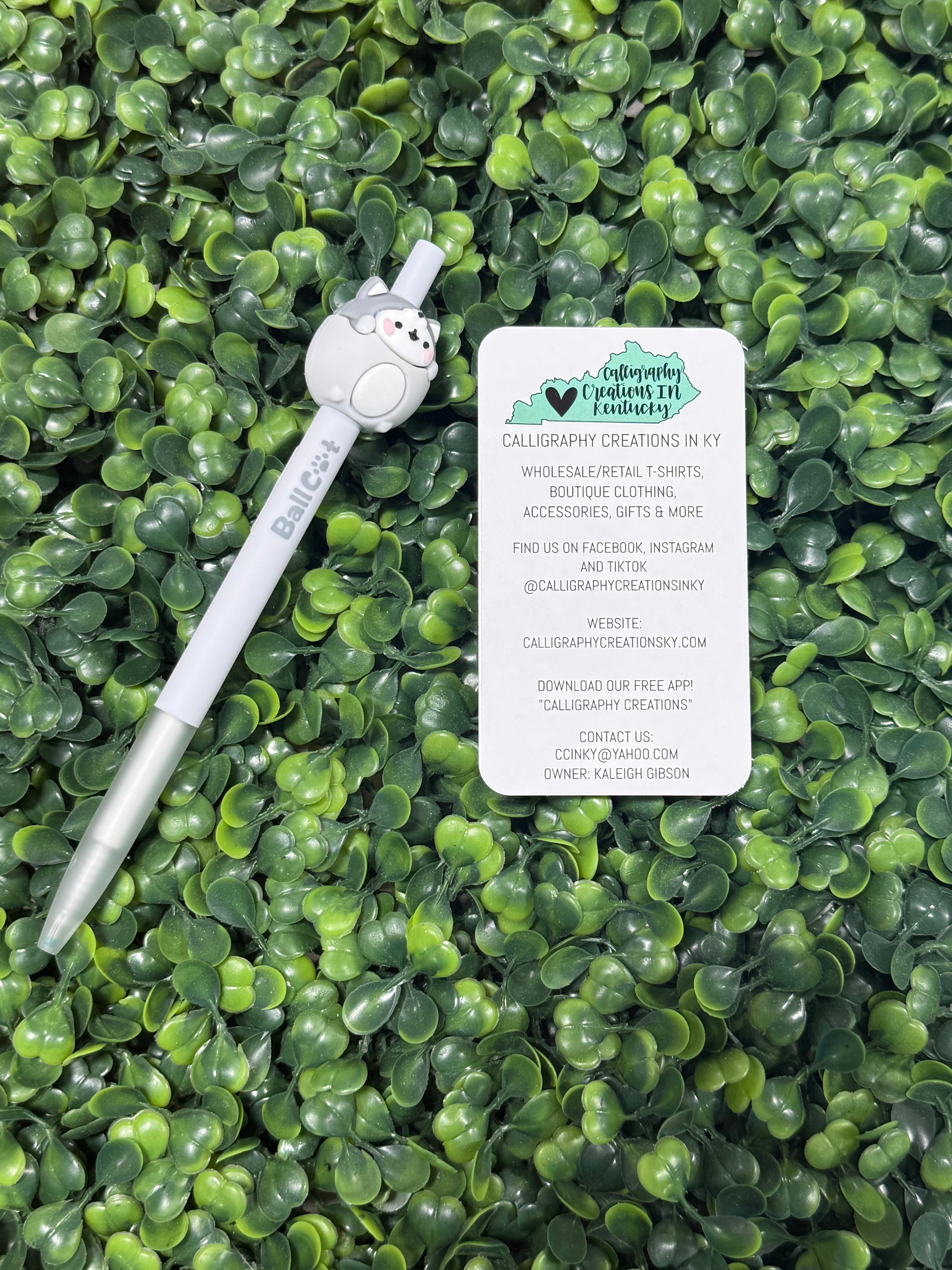 White pen with a small grey cat on top, next to a business card on green leafy background