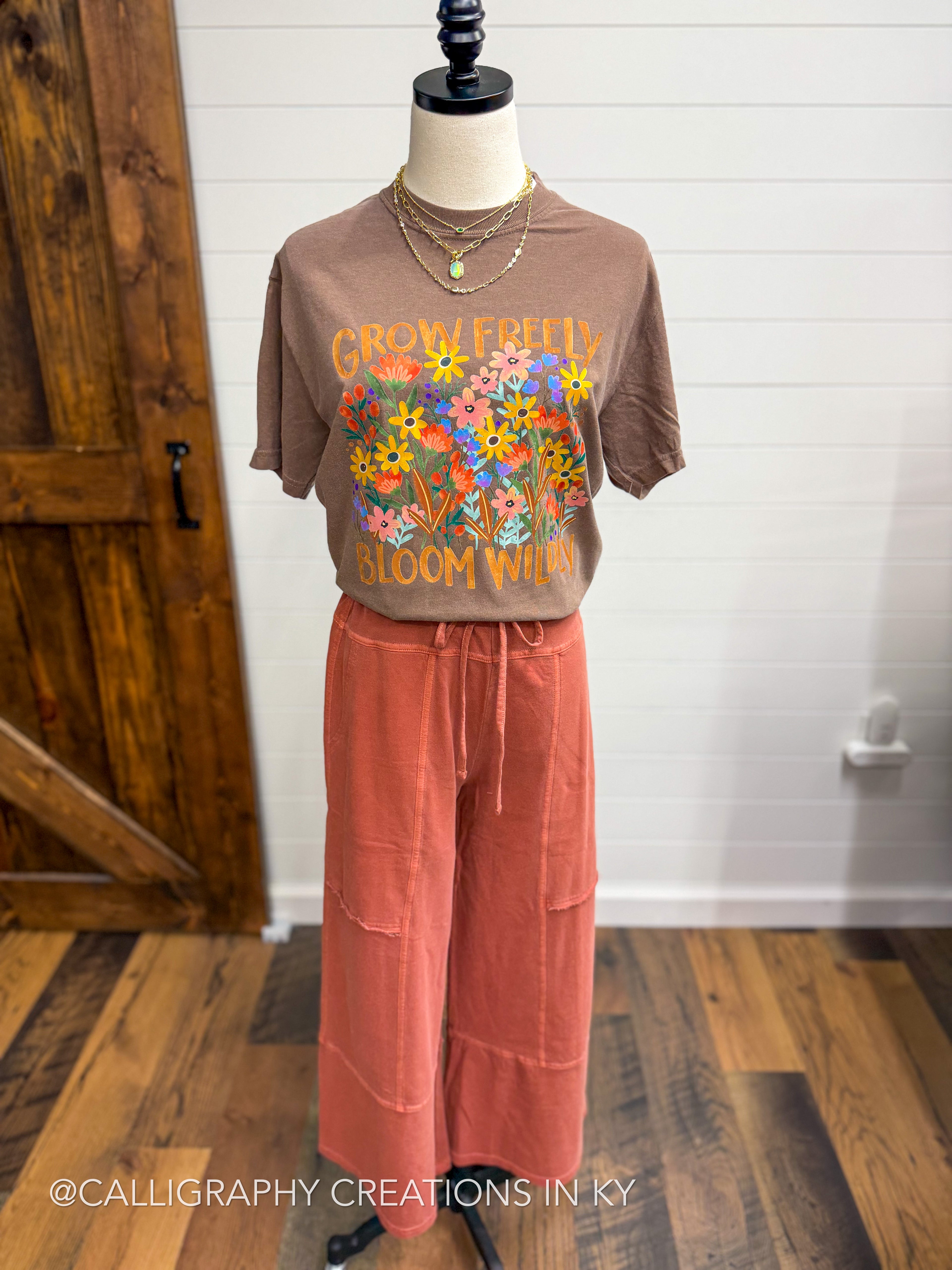 A brown graphic t-shirt with a colorful floral print and the words 'GROW YOURSELF BLOOM WILD' displayed on a mannequin, paired with wide-leg coral pants in a home setting.