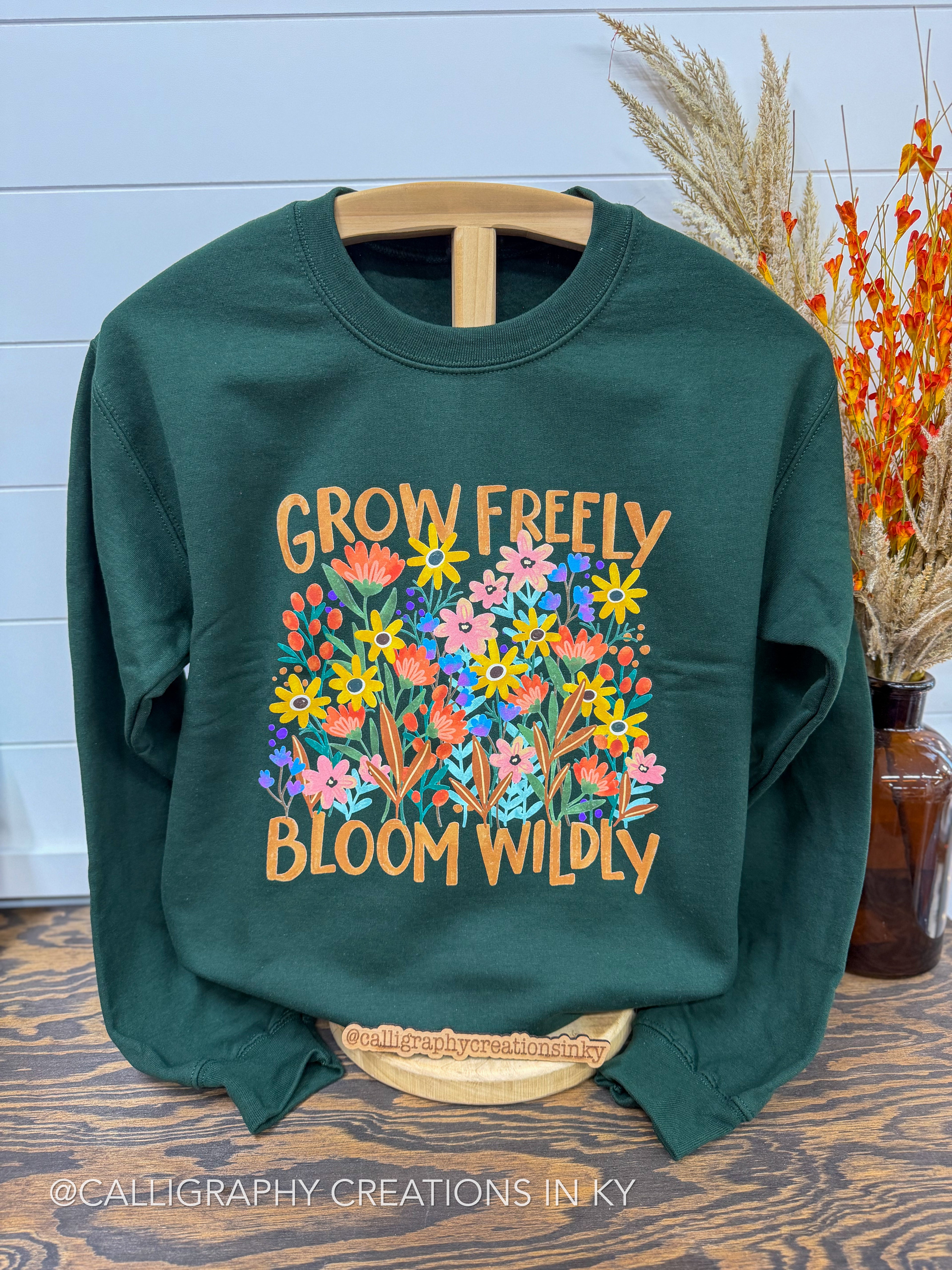 A dark green crewneck sweatshirt with a colorful floral graphic and the text 'GROW FREELY BLOOM WILDLY' in orange, displayed on a wooden hanger on a wooden table with dried grass in a vase in the background.