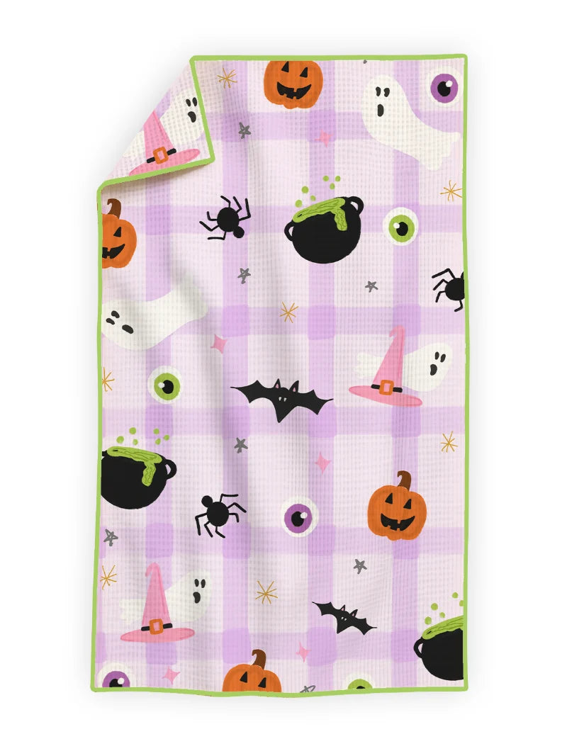 A rectangular kitchen towel with a light purple and white gingham pattern, decorated with a cute Halloween theme including pumpkins, cauldrons, spiders, ghosts, and bats. The blanket features a solid light green border and is shown with one corner folded over, set against a plain white background.