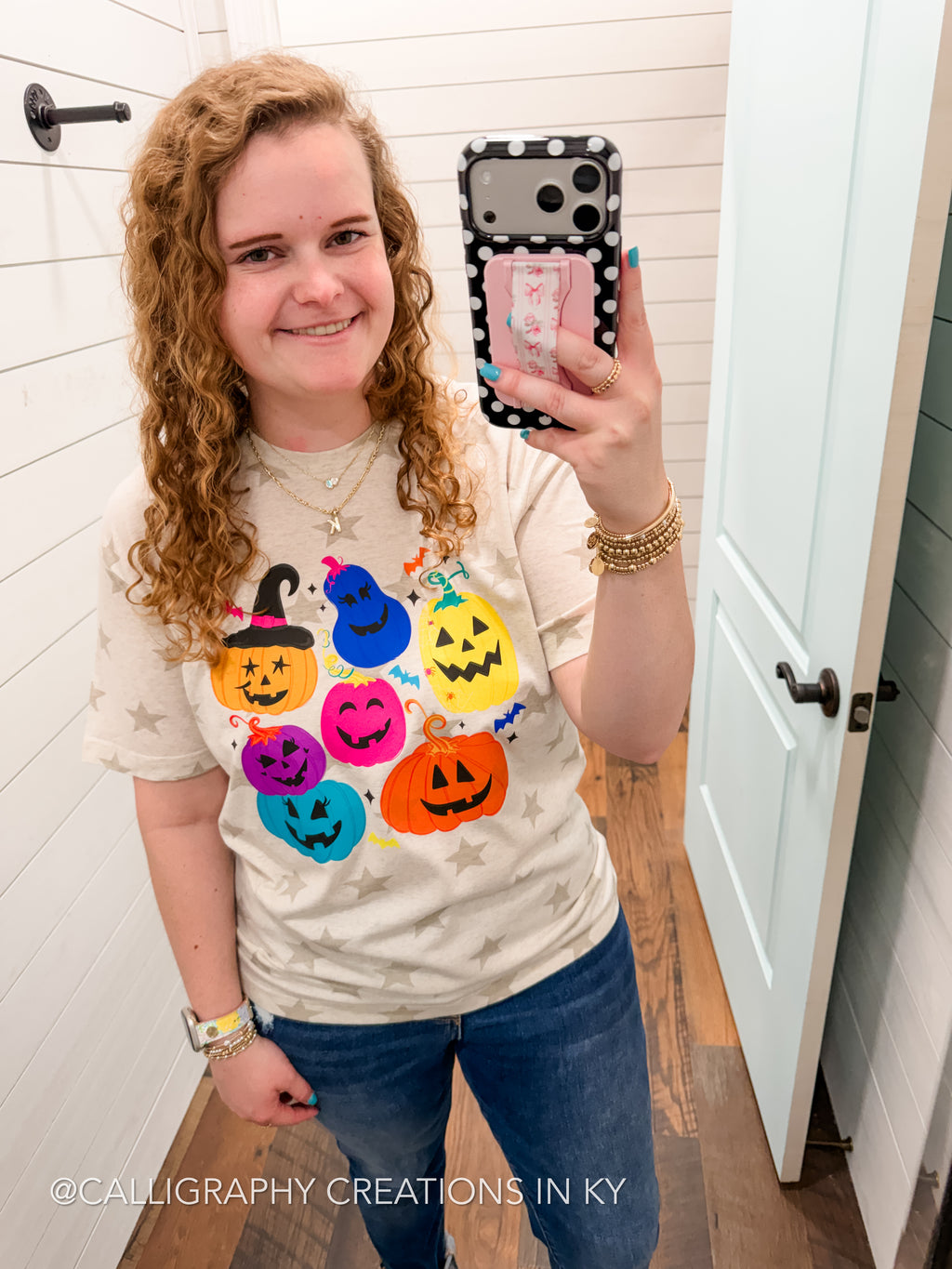 A woman takes a mirror selfie wearing a cream-colored t-shirt with a colorful graphic of jack-o'-lanterns and ghosts, paired with blue jeans. The watermark at the bottom reads '@CALLIGRAPHY CREATIONS IN KY'.