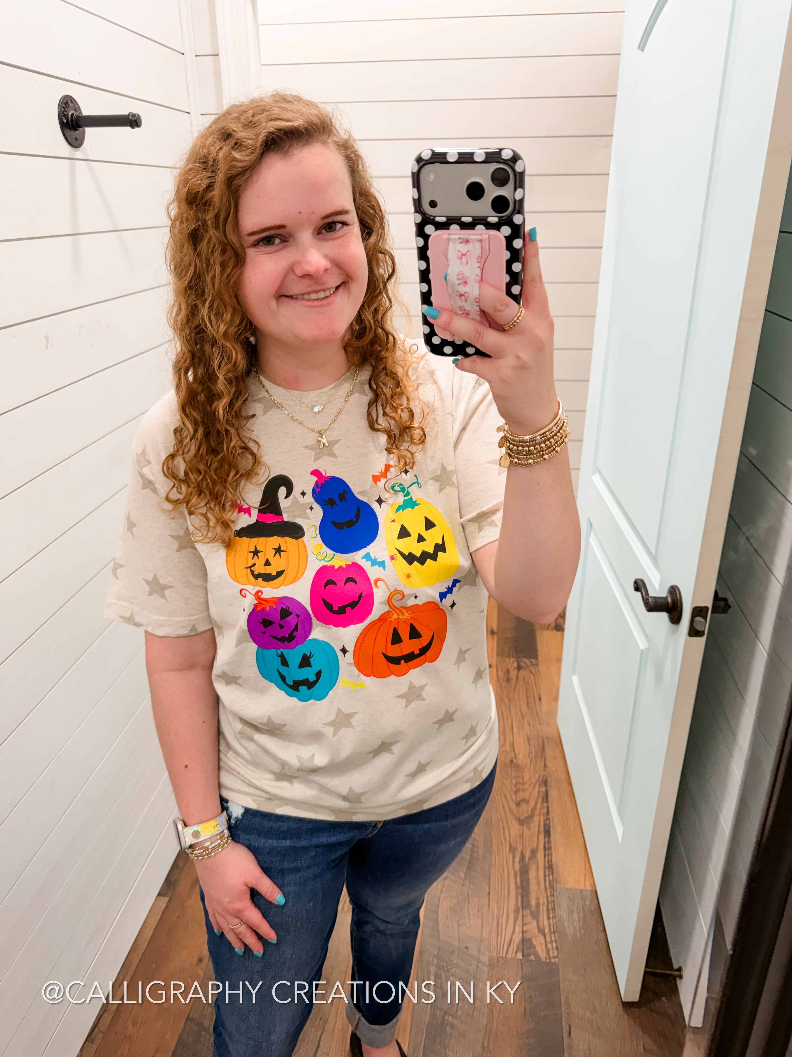 A woman taking a mirror selfie wearing a beige graphic t-shirt with colorful jack-o'-lanterns and stars, paired with blue jeans.