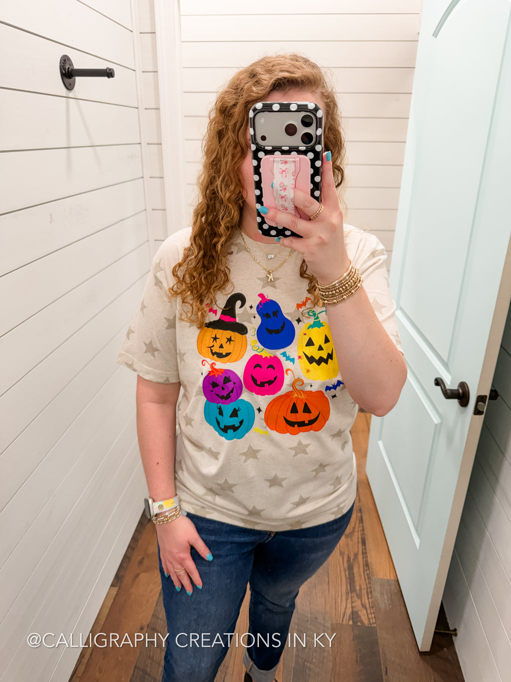 A woman takes a mirror selfie in a fitting room wearing a beige t-shirt with a colorful graphic of jack-o'-lanterns in different colors, paired with blue jeans.