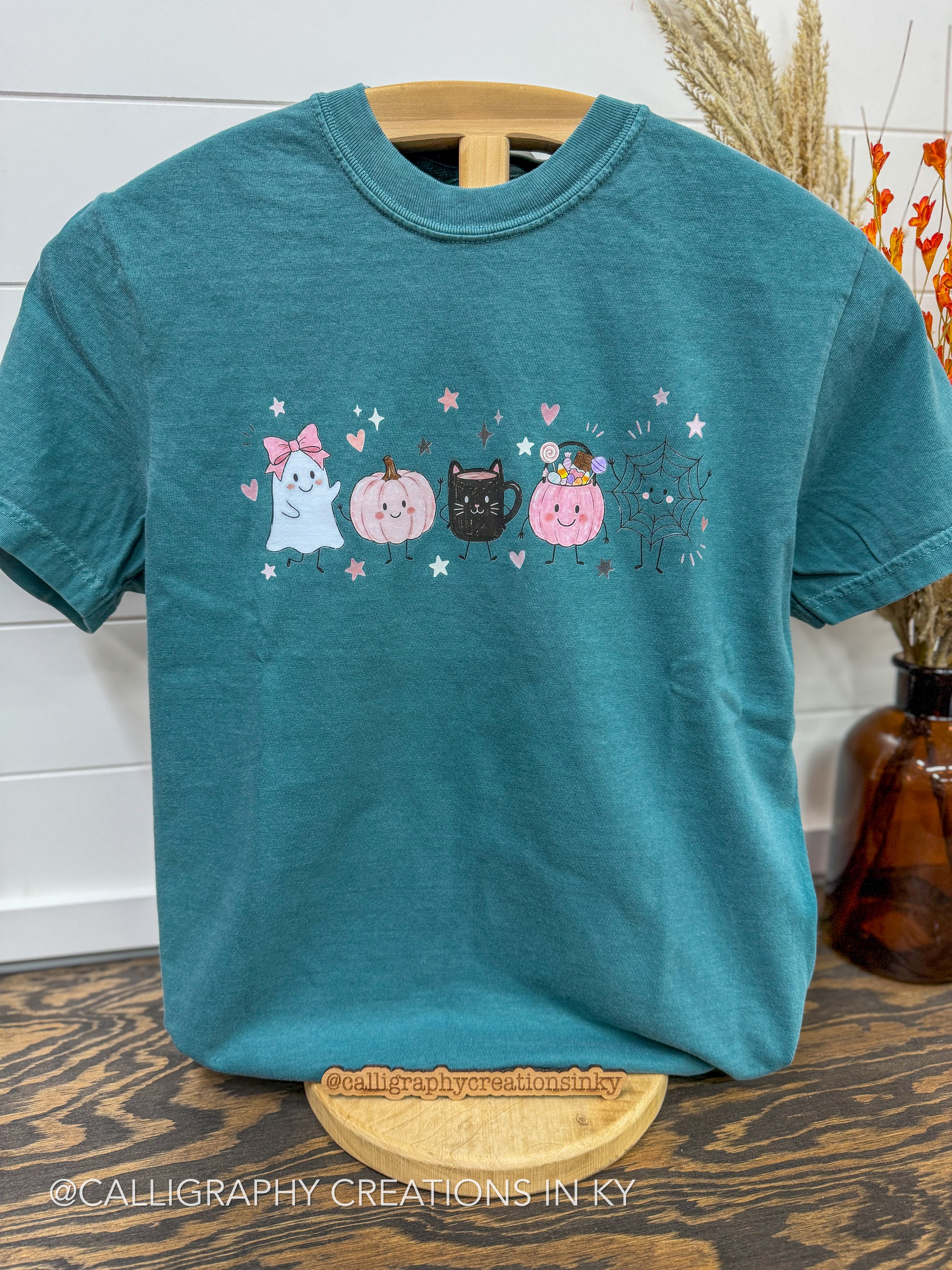 A teal short-sleeve t-shirt on a wooden hanger, featuring a cute graphic of four cats dressed as pumpkins (ghost, orange pumpkin, black cat, pink witch pumpkin) with stars and hearts. The shirt is displayed on a wooden stand on a dark wood table with a white shiplap wall and dried grass in the background.