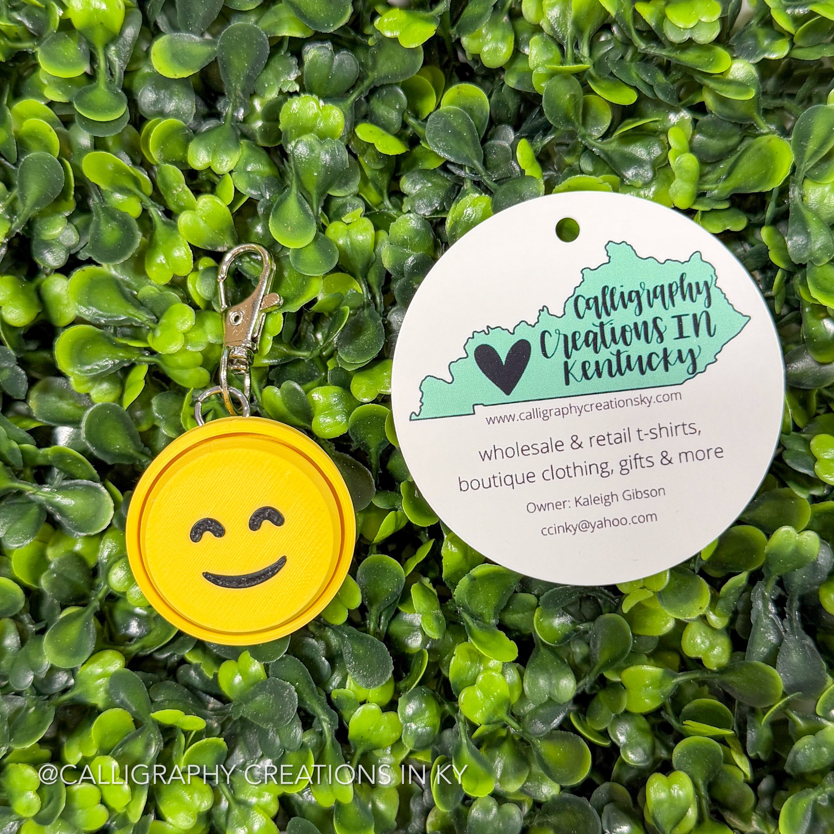 Yellow keychain with happy face on greenery background with Calligraphy Creation branding