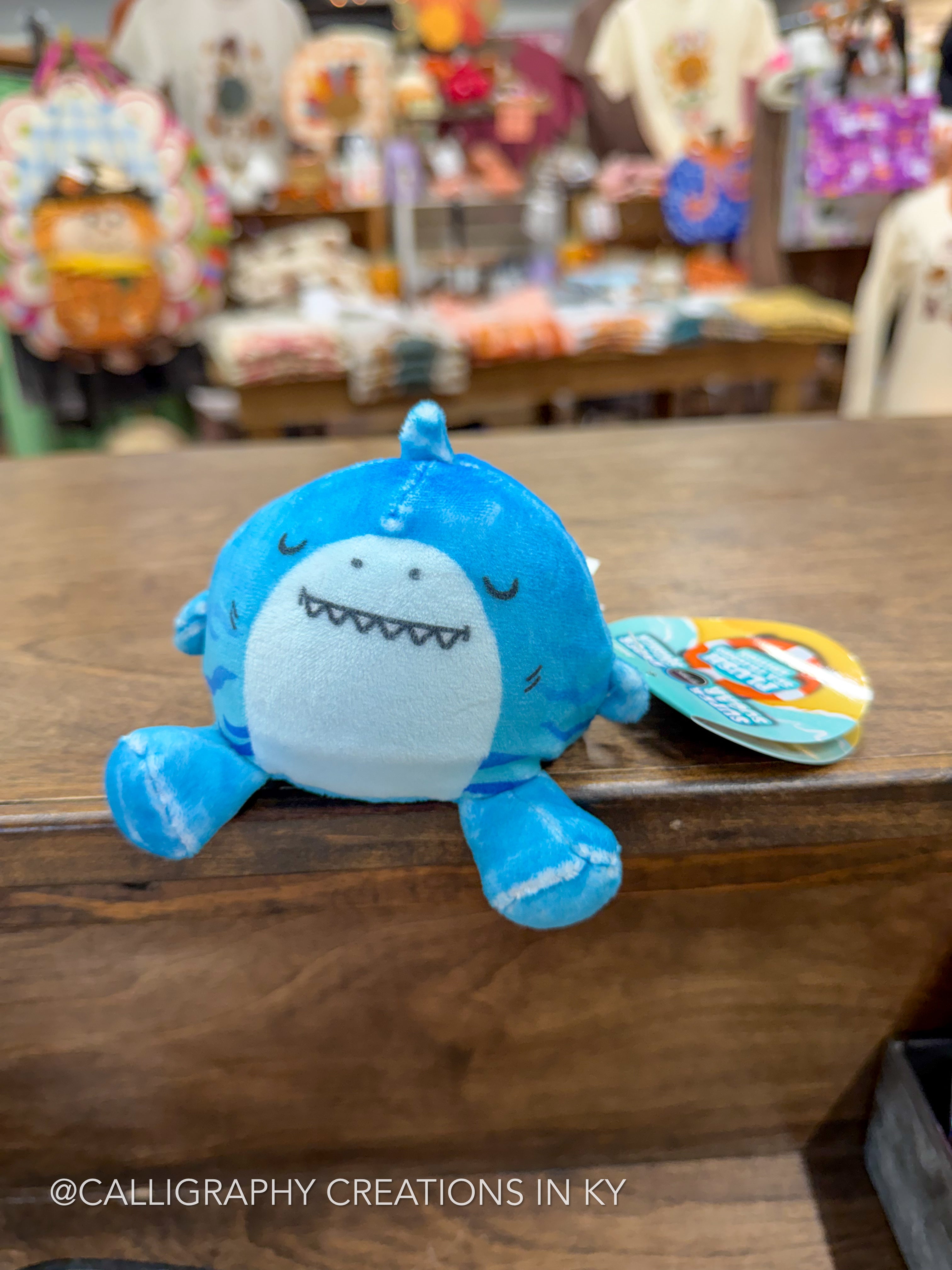 A blue and white plush shark toy with a stitched smile and closed eyes, resting on a wooden counter in a store setting with a colorful tag attached.