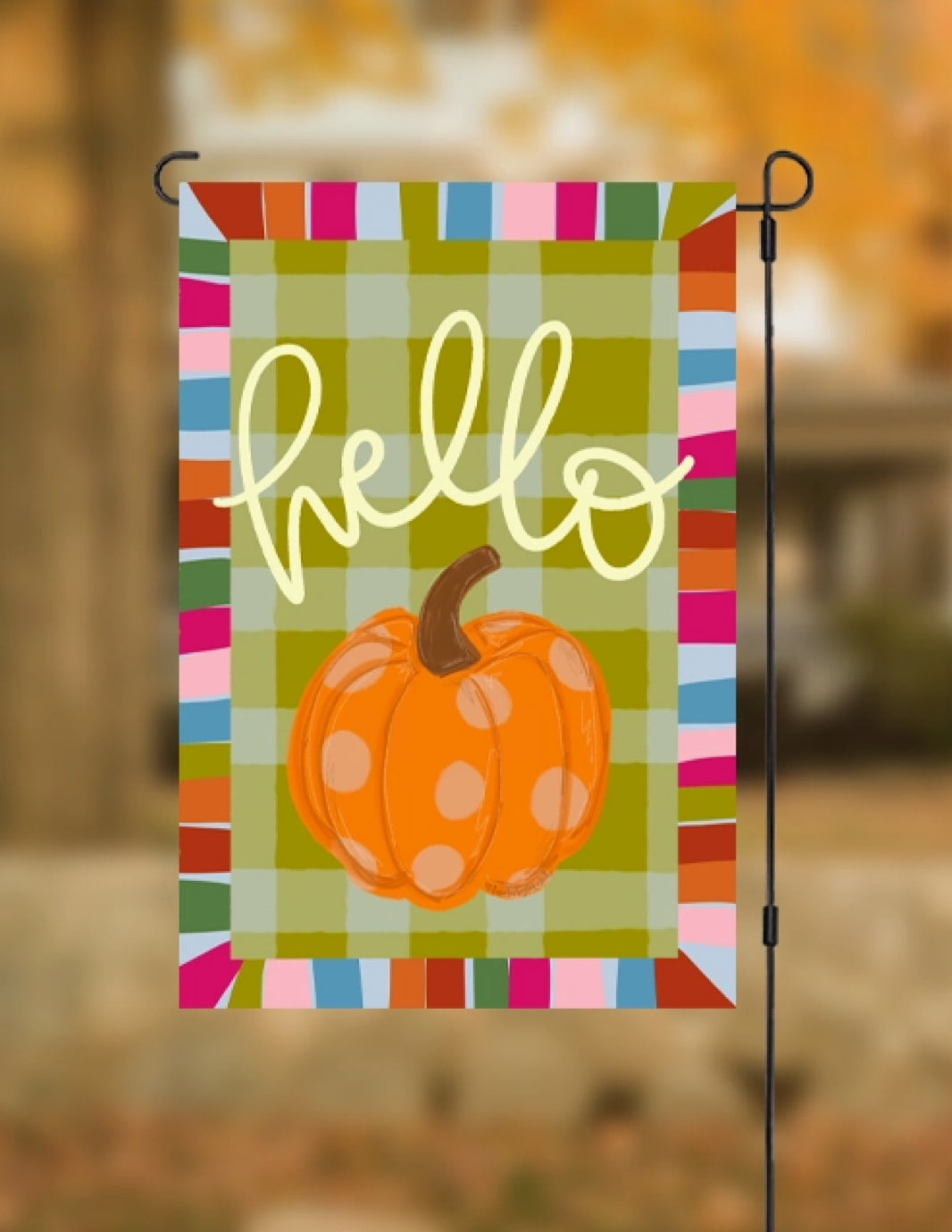 A vertical garden flag with a colorful striped border, olive green plaid background, yellow 'hello' script, and an orange pumpkin with white polka dots, shown hanging on a black metal stand against a blurred autumn outdoor background.