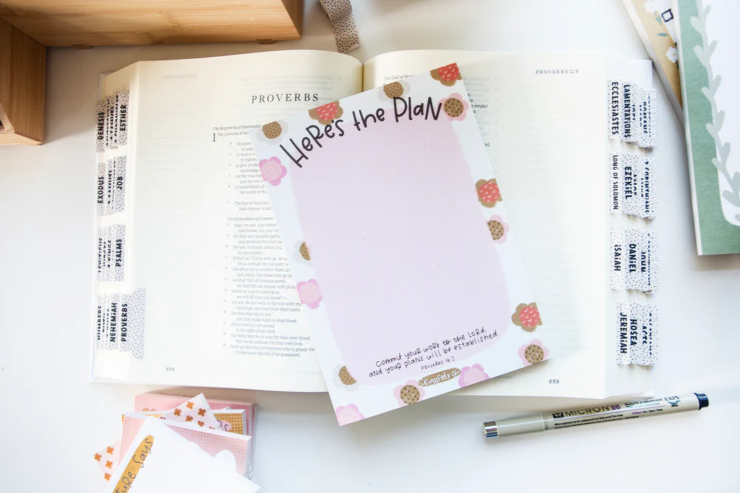 A styled flat-lay of a planner notepad with the title 'Here's the Plan' resting on an open Bible, surrounded by planner accessories including sticky notes, a pen, and washi tape on a white surface.