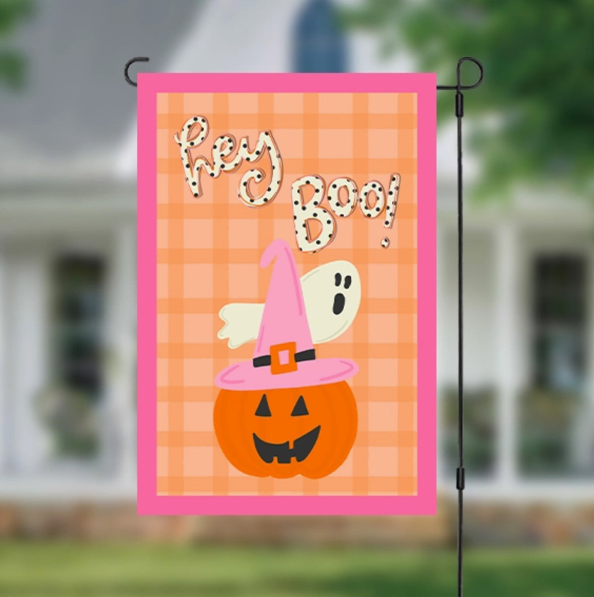 A Halloween garden flag hanging on a black metal stand, featuring a pink border, orange plaid background, a jack-o'-lantern, a ghost, and the words 'Hey Boo!' in a spooky font. The background shows a blurred house and lawn.