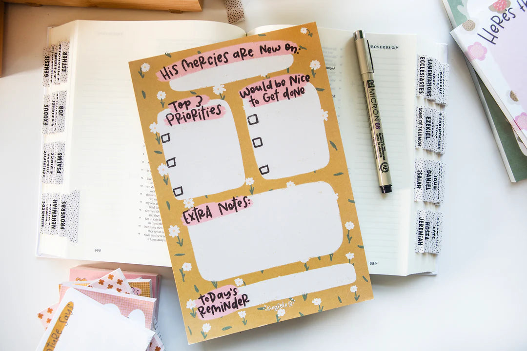 A decorative daily planner insert sheet with floral doodles and handwritten notes, placed on top of an open notebook on a light tabletop. A pen, additional tabs, and other stationery items are visible around it.