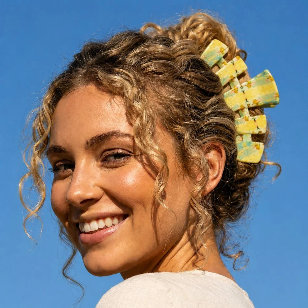 A smiling woman with curly blonde hair in an updo wearing a large yellow and green marbled claw hair clip, set against a clear blue sky.