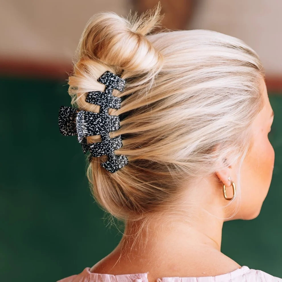 A close-up side view of a woman with blonde hair styled in a messy bun, secured with a large black claw hair clip She is wearing a small gold hoop earring, and the background is a soft, out-of-focus dark green.