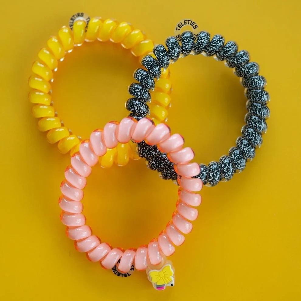 Three coiled elastic hair ties in yellow, pink, and blue-gray are arranged on a bright yellow background. The pink one has a small butterfly charm attached.