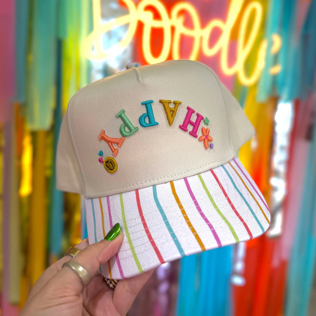 Beige cap with colorful 'Happy' text and striped brim held in front of a vibrant, multicolored background.