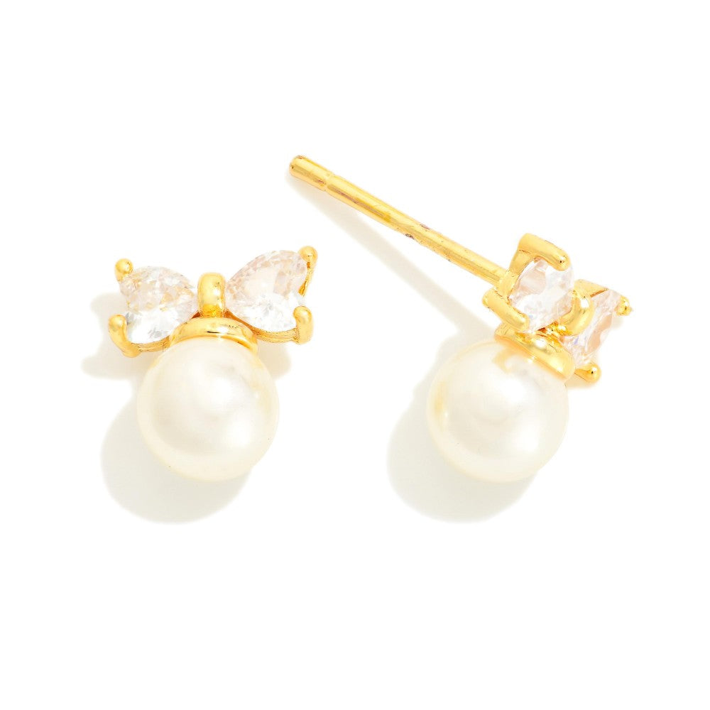 A pair of gold stud earrings featuring a small bow-like cluster of clear marquise-cut stones above a round white pearl, shown on a clean white background.