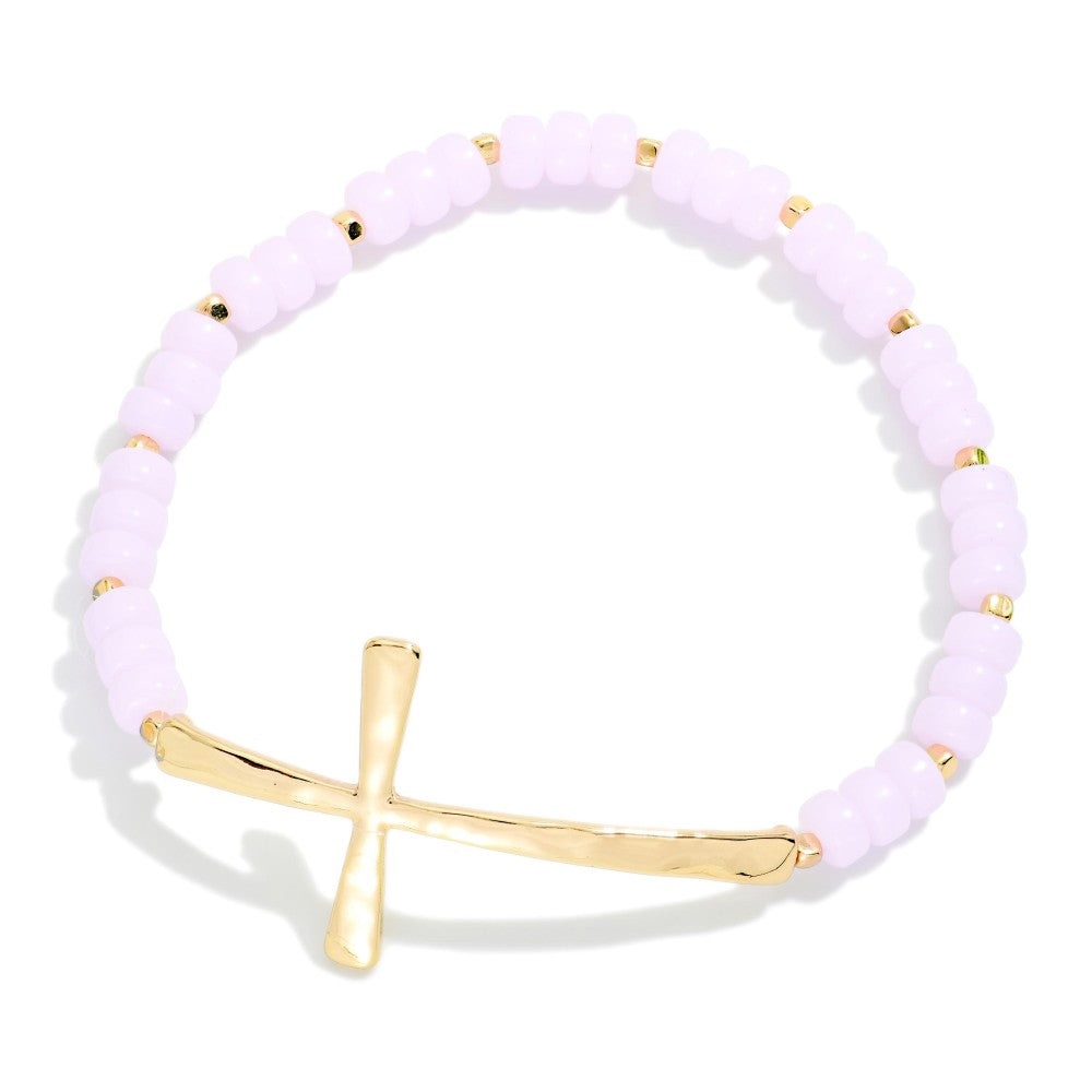 A beaded stretch bracelet featuring pale lavender beads separated by small gold spacer beads, with a polished gold cross charm at the front, set against a clean white background.