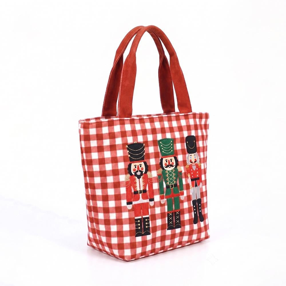 A red and white gingham canvas tote bag with brown fabric handles, featuring a printed graphic of three nutcracker figures in red, green, and black uniforms.