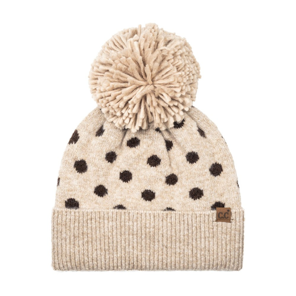 A beige knit pom-pom beanie with dark brown polka dots and a ribbed cuff, featuring a small brown leather patch with 'CG' on the side.