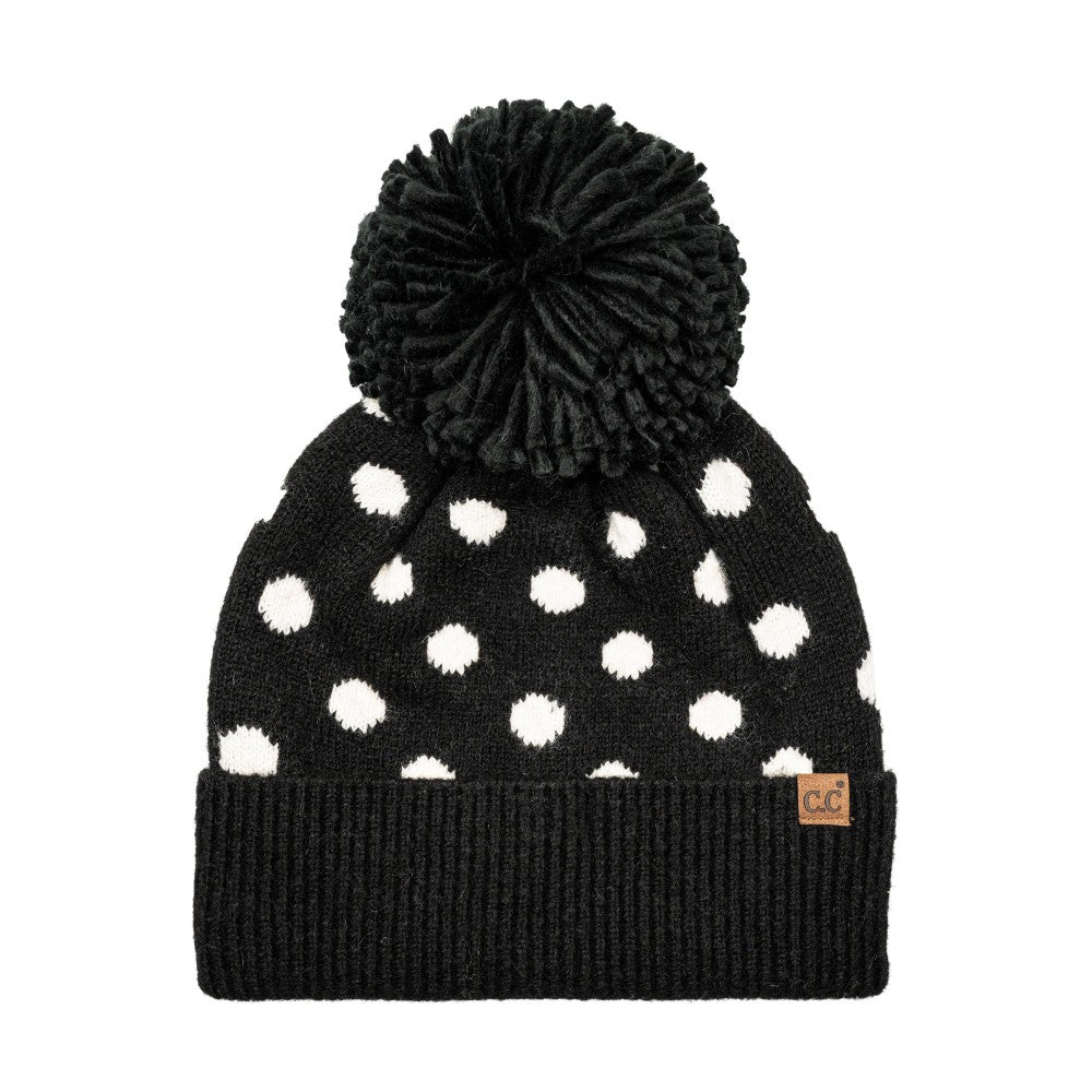 A black knit beanie with white polka dots and a large black pom-pom on top, featuring a ribbed cuff and a small tan square logo patch on the side.