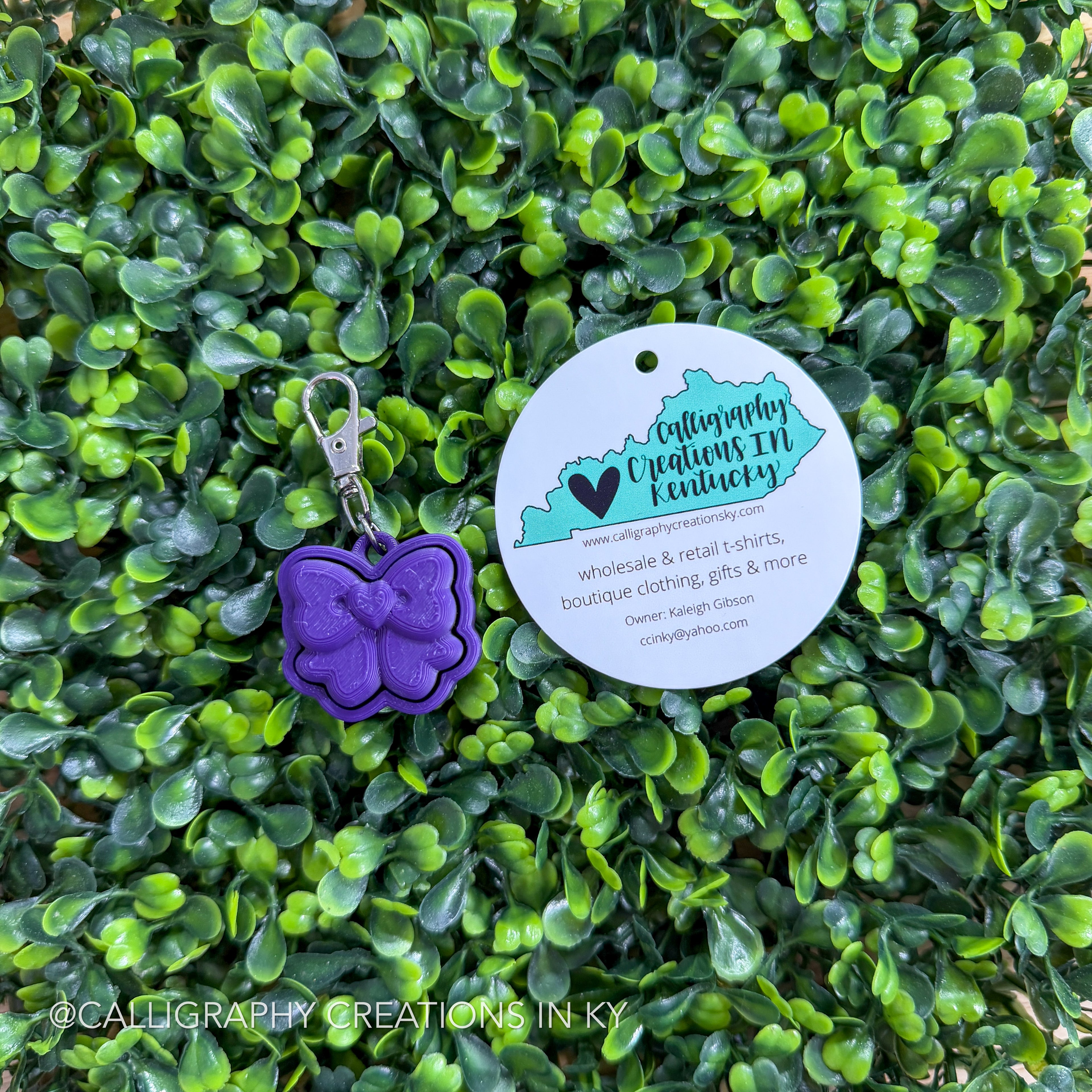 Purple bow shaped fidget clicker keychain with a tag on a green leafy background