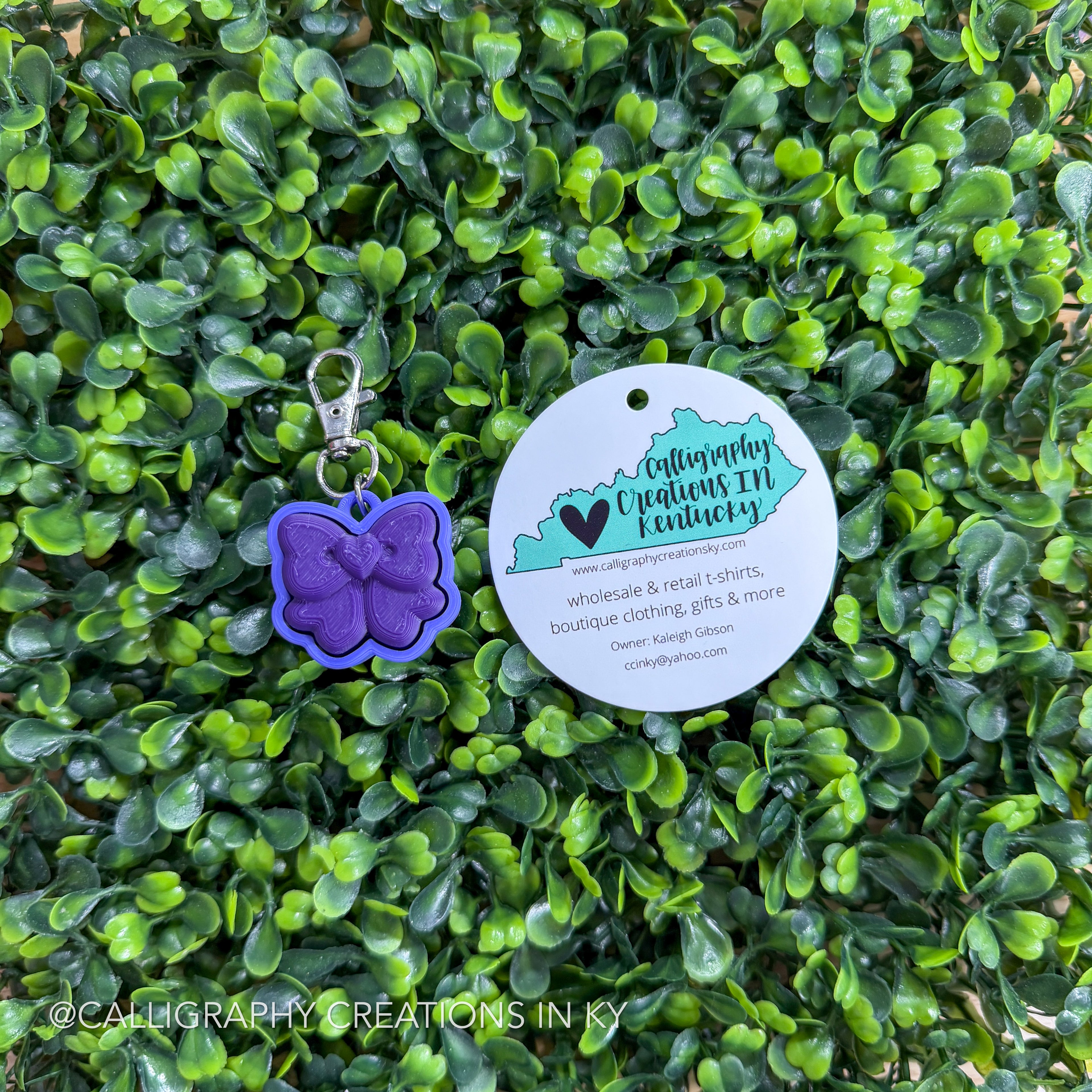 Purple bow fidget clicker with a round tag on green Leafy background, featuring Calligraphy Creations in KY branding.