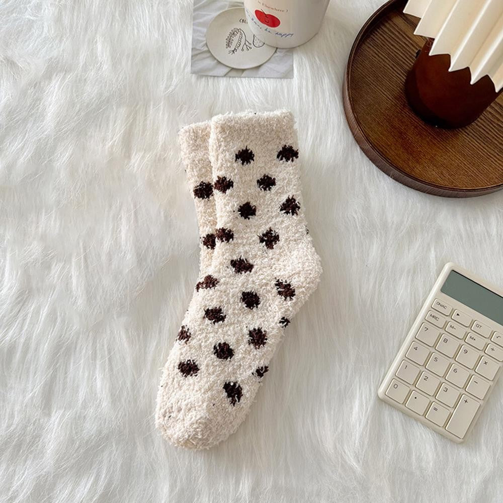 A pair of cream-colored fuzzy socks with dark brown polka dots, styled on a white faux fur surface with a wooden tray, a calculator, and a cup in the background.