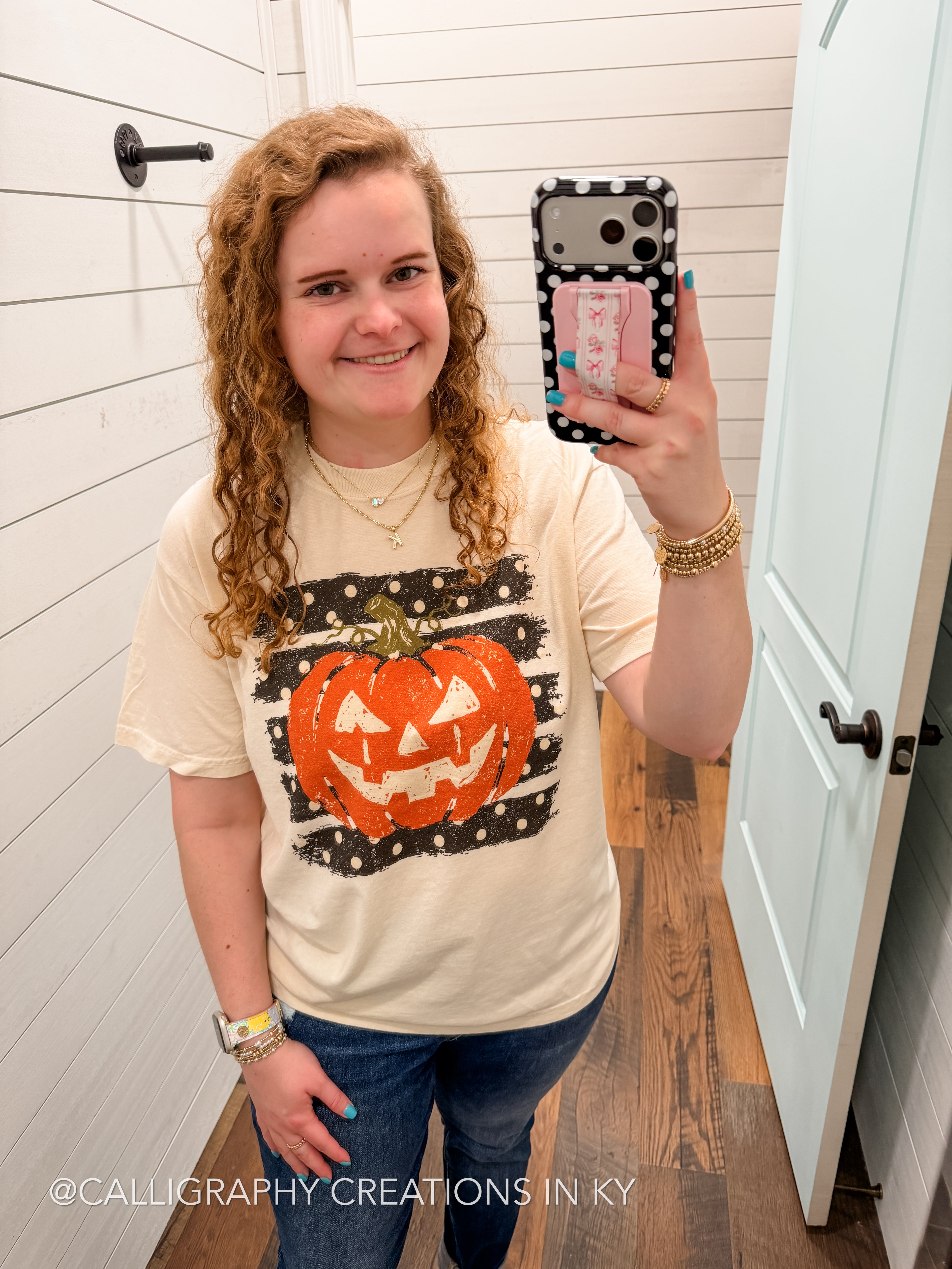 A woman takes a mirror selfie in a bathroom wearing a cream-colored t-shirt with a distressed black and white pattern featuring an orange jack-o'-lantern graphic. She is also wearing blue jeans and holding a phone with a polka-dot case.