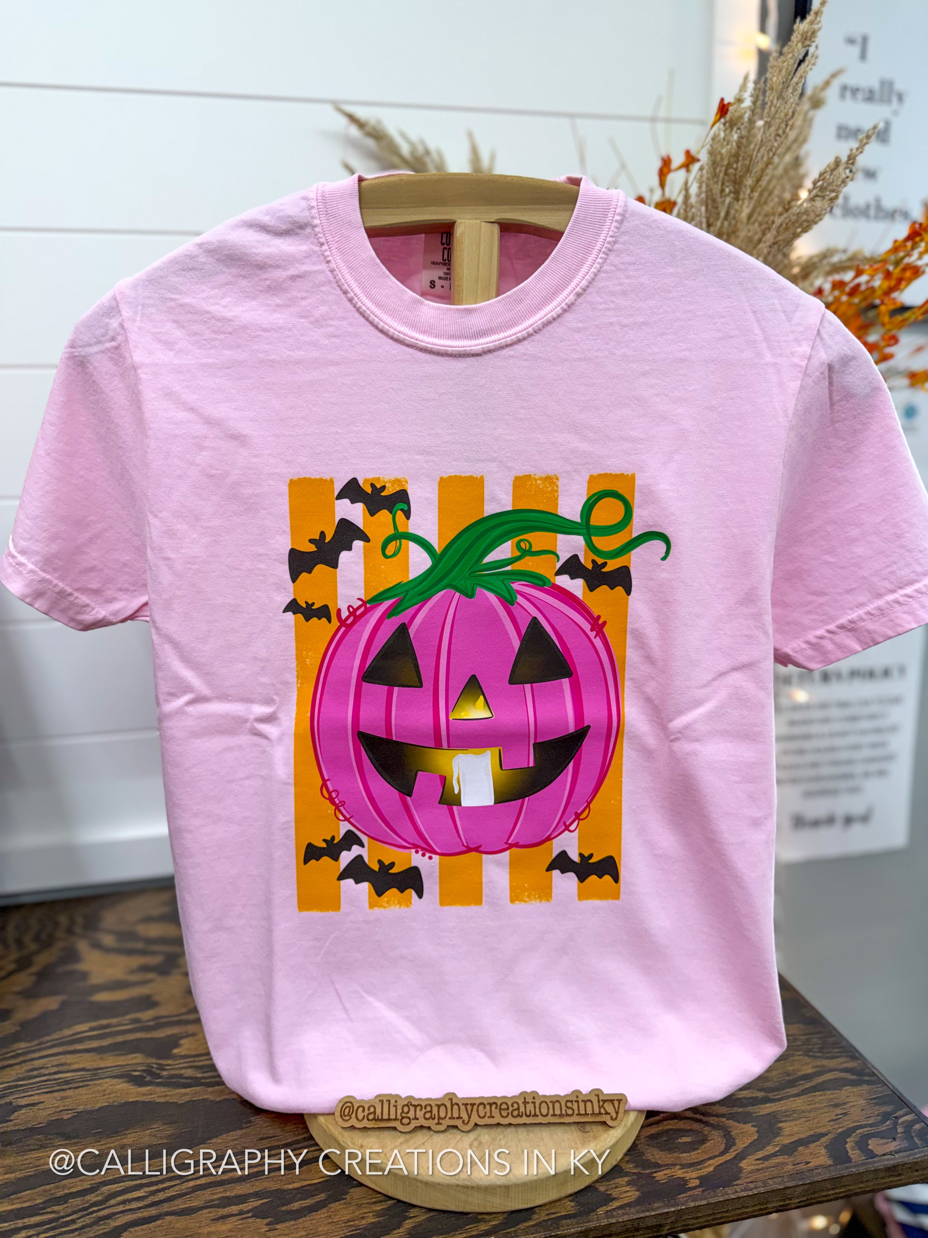 A light pink short-sleeve t-shirt on a wooden hanger, featuring a graphic of a jack-o'-lantern with a pink striped pumpkin body, green stem, and black bat silhouettes on an orange striped background.
