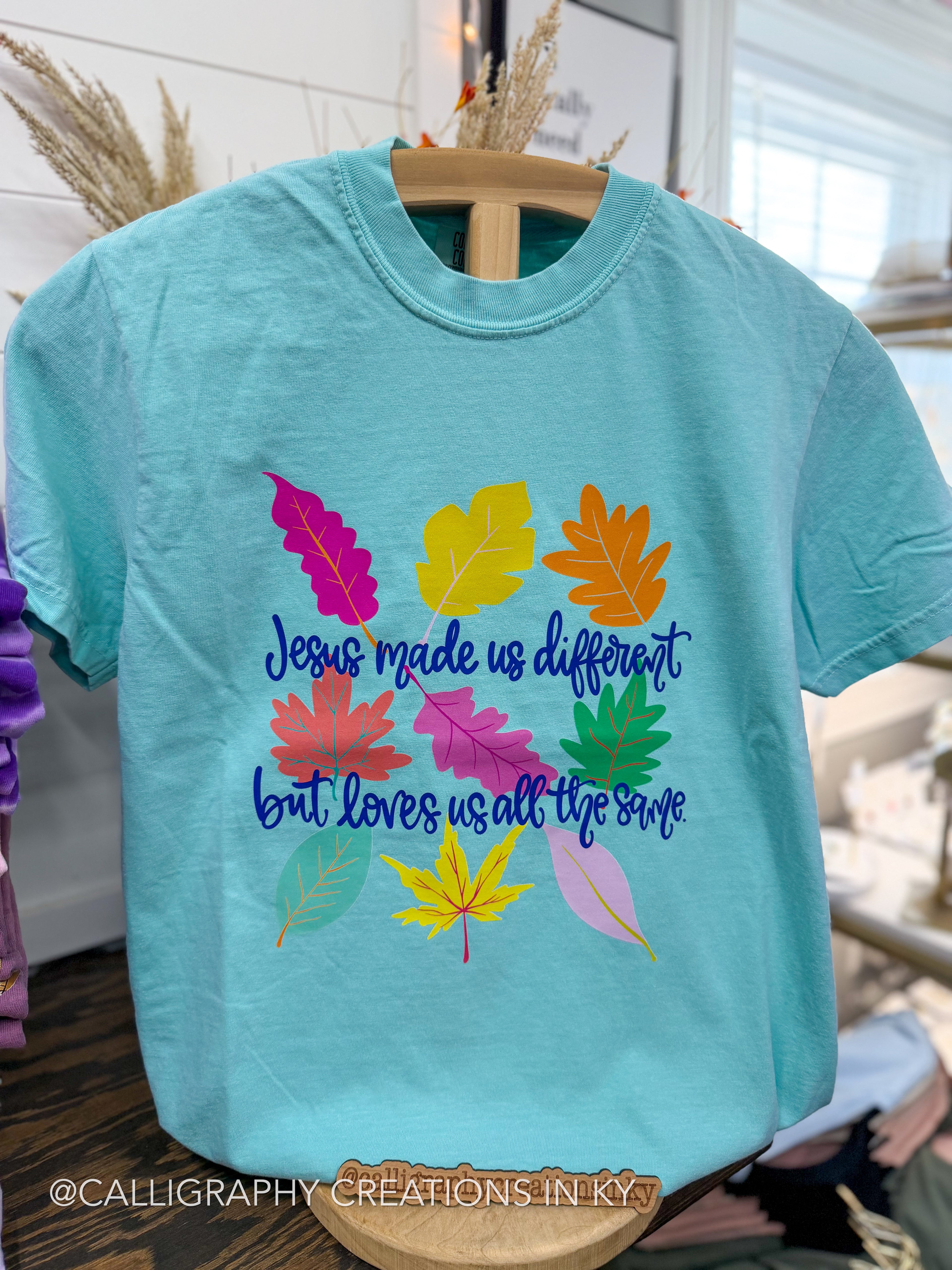 A light blue crewneck t-shirt on a wooden hanger, featuring a colorful graphic of autumn leaves and the text 'Jesus made us different but loves us all the same' in blue cursive. The photo is taken in a retail store setting.