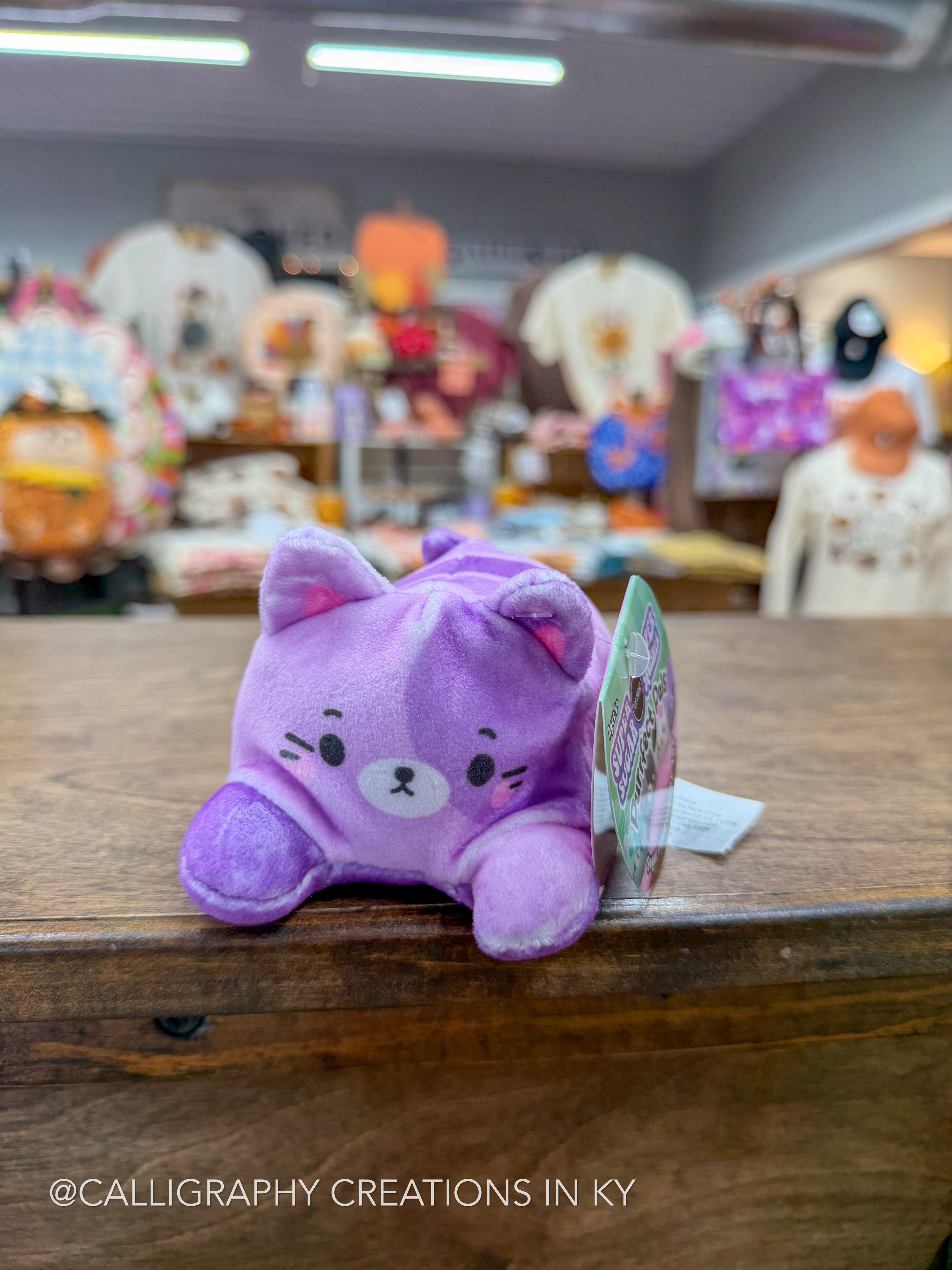 A small purple plush cat toy with embroidered facial features sits on a wooden counter in a store, with a hangtag attached and a blurred shop interior in the background.