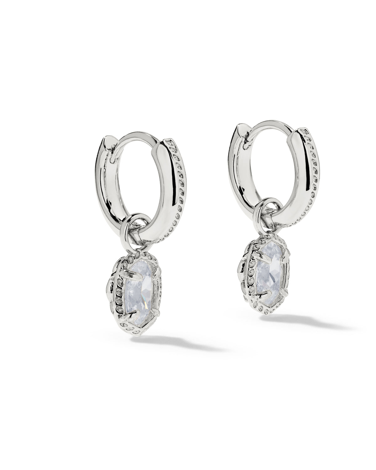 A pair of silver huggie hoop earrings with dangling oval clear gemstones, set against a plain white background.