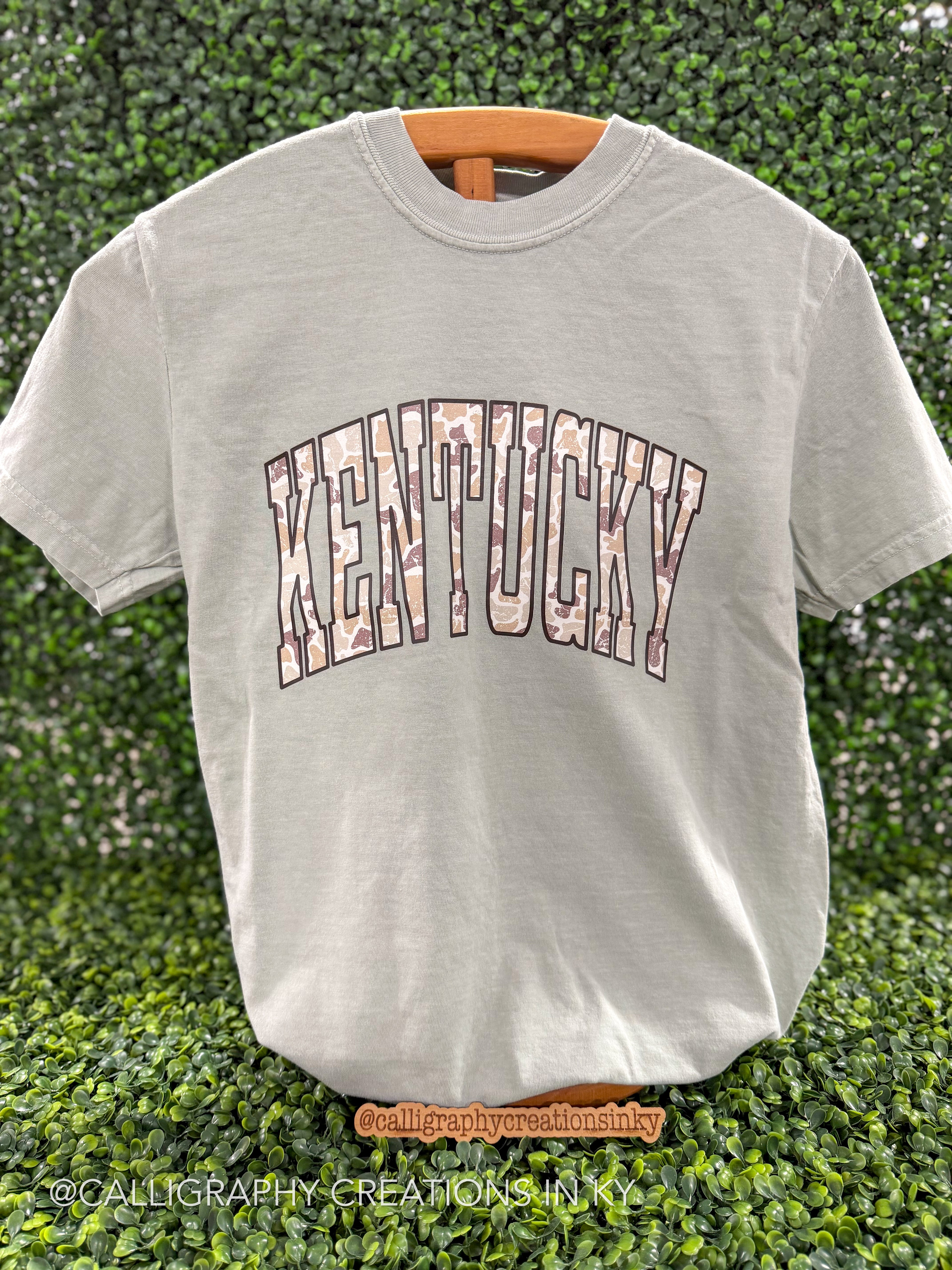 Sage green shirt with Camo ‘Kentucky’ across the chest on a wooden stand