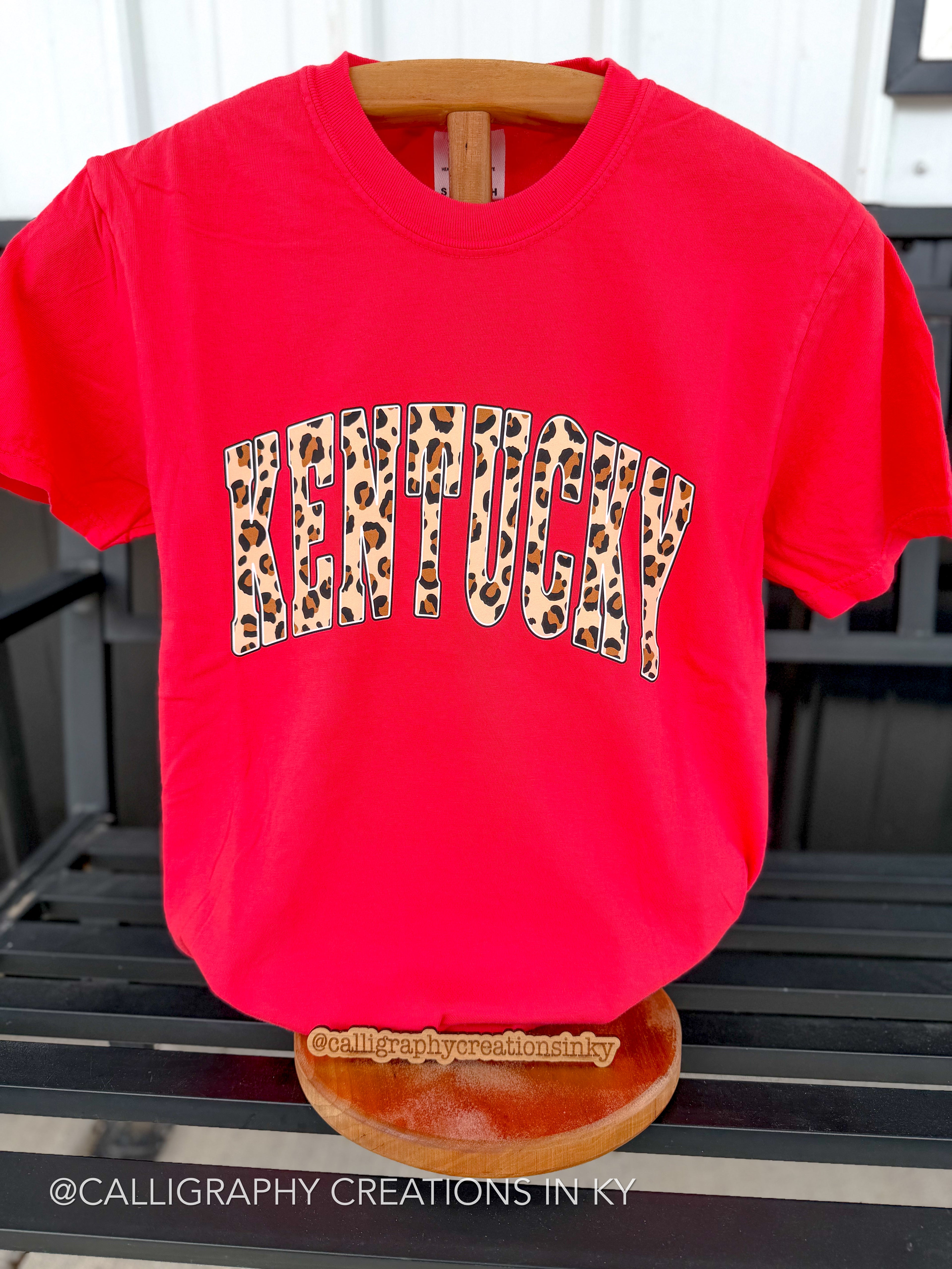 A bright red short-sleeve t-shirt displayed on a wooden hanger and stand. The front features the word 'KENTUCKY' in an arched leopard-print pattern.
