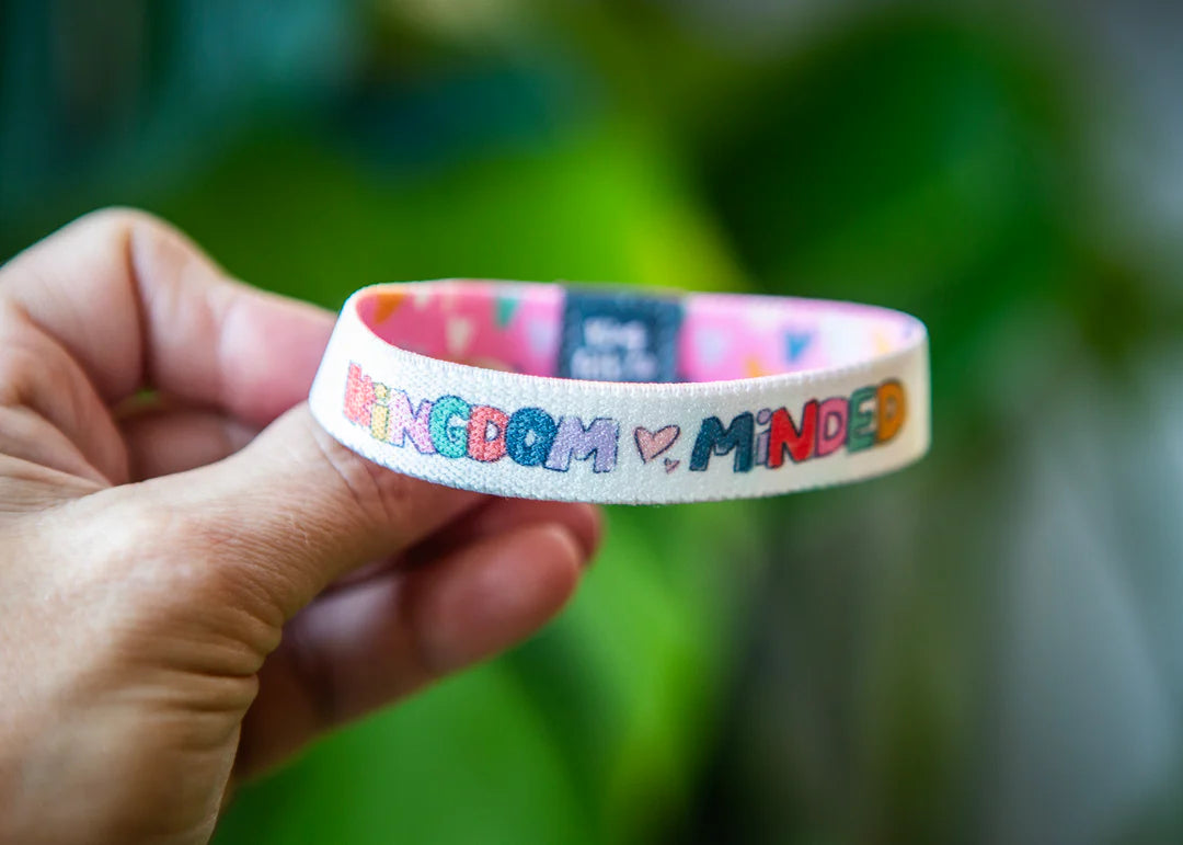 A hand holding a white fabric wristband with colorful embroidered text reading 'Kingdom Minded' and a small heart in the center, against a blurred green background.