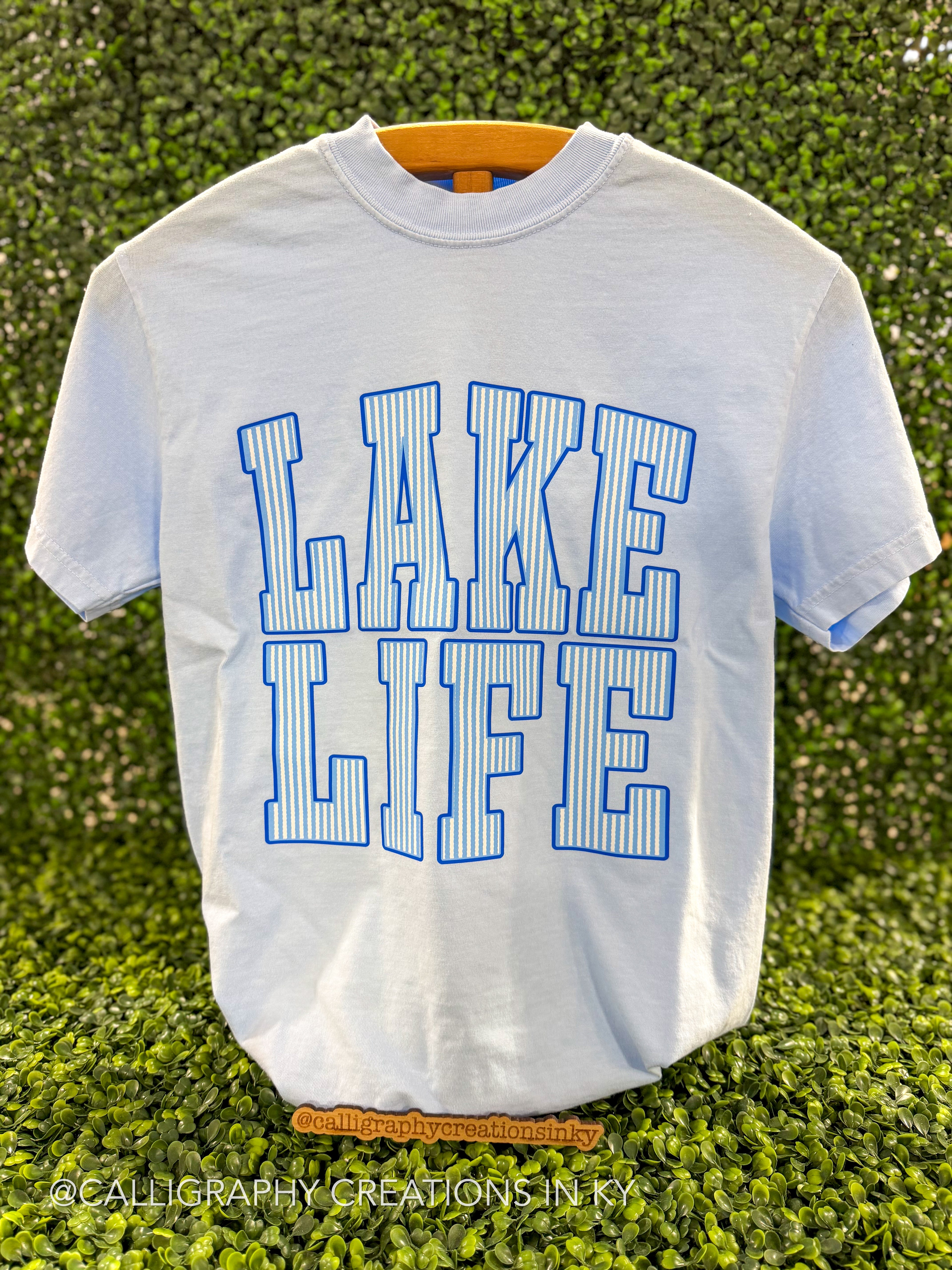 Blue shirt with blue and white striped letters spelling ‘Lake Life’ on a wooden stand 