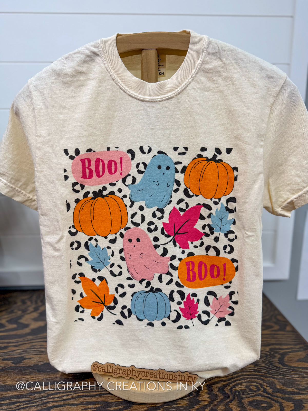 A cream-colored short-sleeve t-shirt with a colorful autumn-themed graphic print featuring pumpkins, ghosts, leaves, and 'BOO!' text on a black leopard print background, displayed on a wooden hanger.