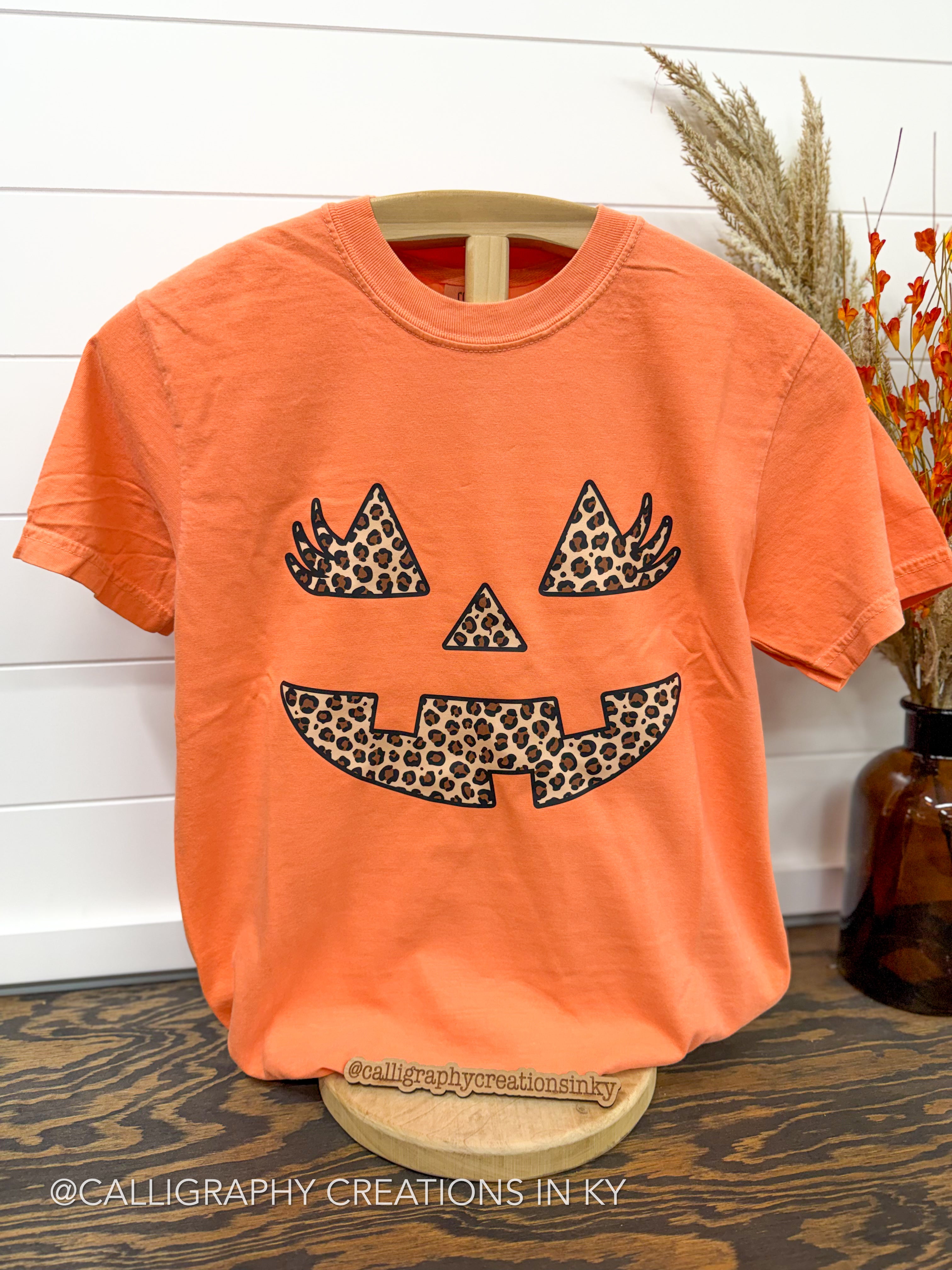 An orange short-sleeve t-shirt displayed on a wooden hanger, featuring a jack-o'-lantern face graphic filled with a black and tan leopard print pattern. The shirt is set against a white shiplap wall with dried grasses and an amber glass vase on a dark wood floor.