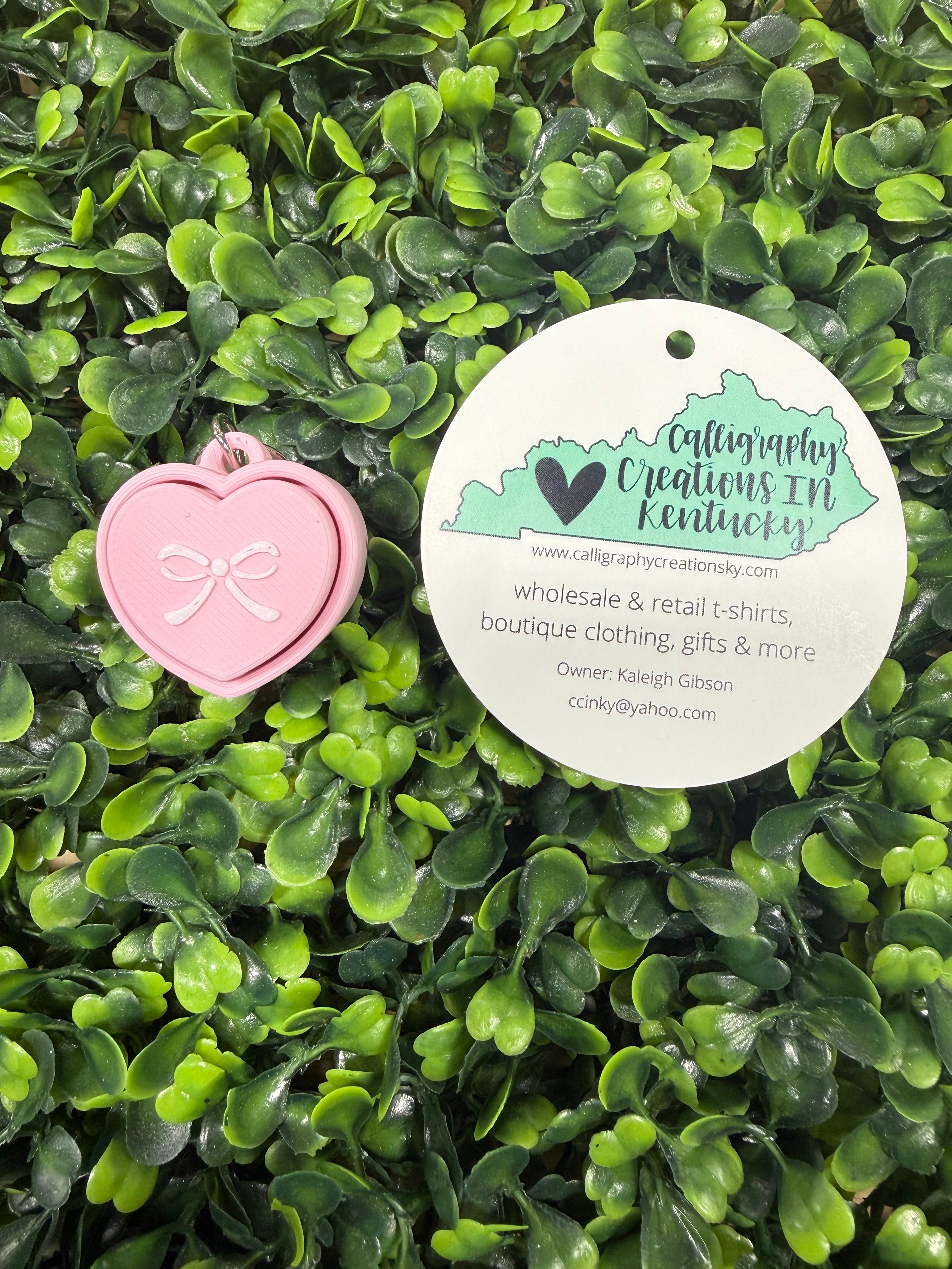 Pink heart-shaped keychain with a label on a bed of green leaves