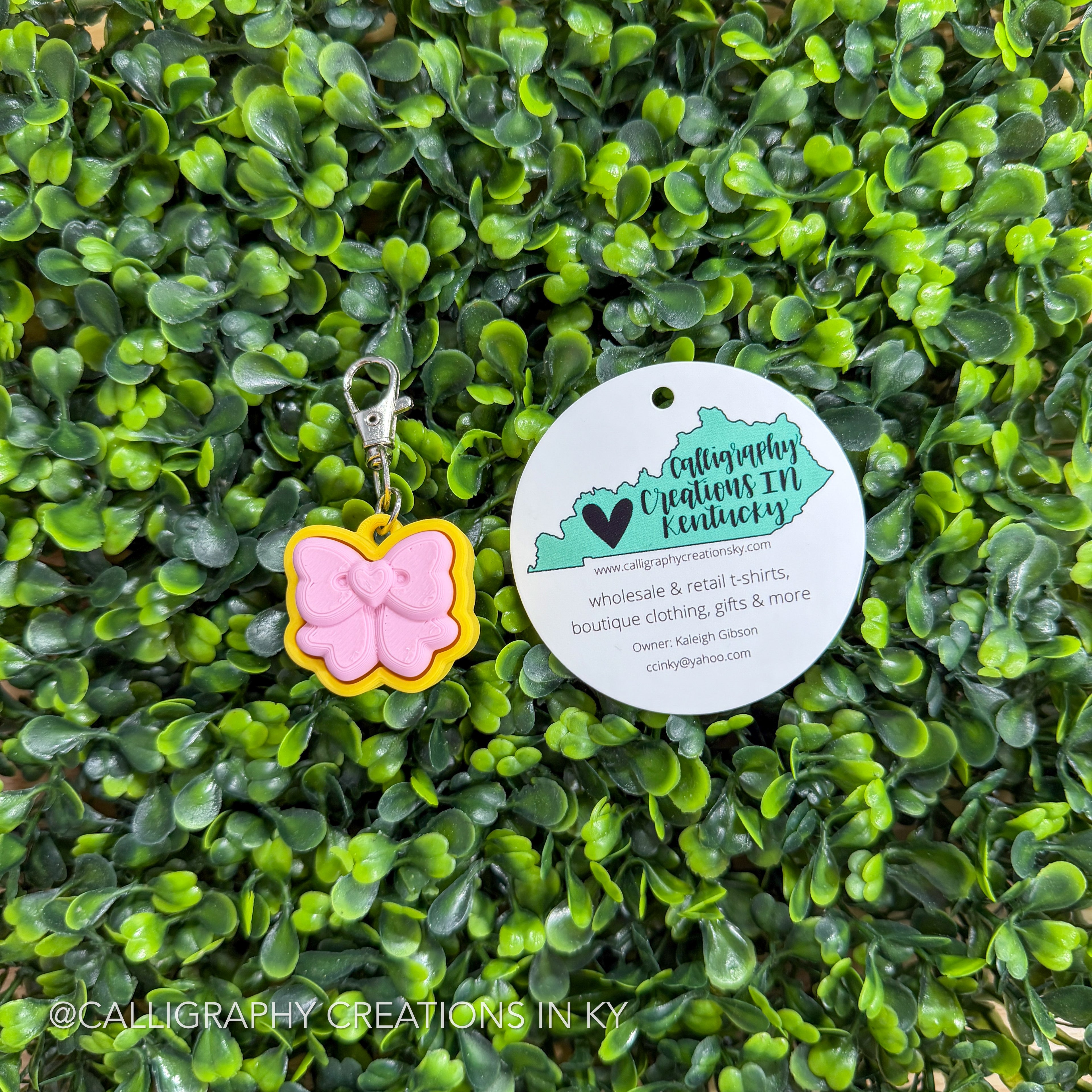 Pink and yellow bow fidget clicker keychain with a tag on a green leafy background