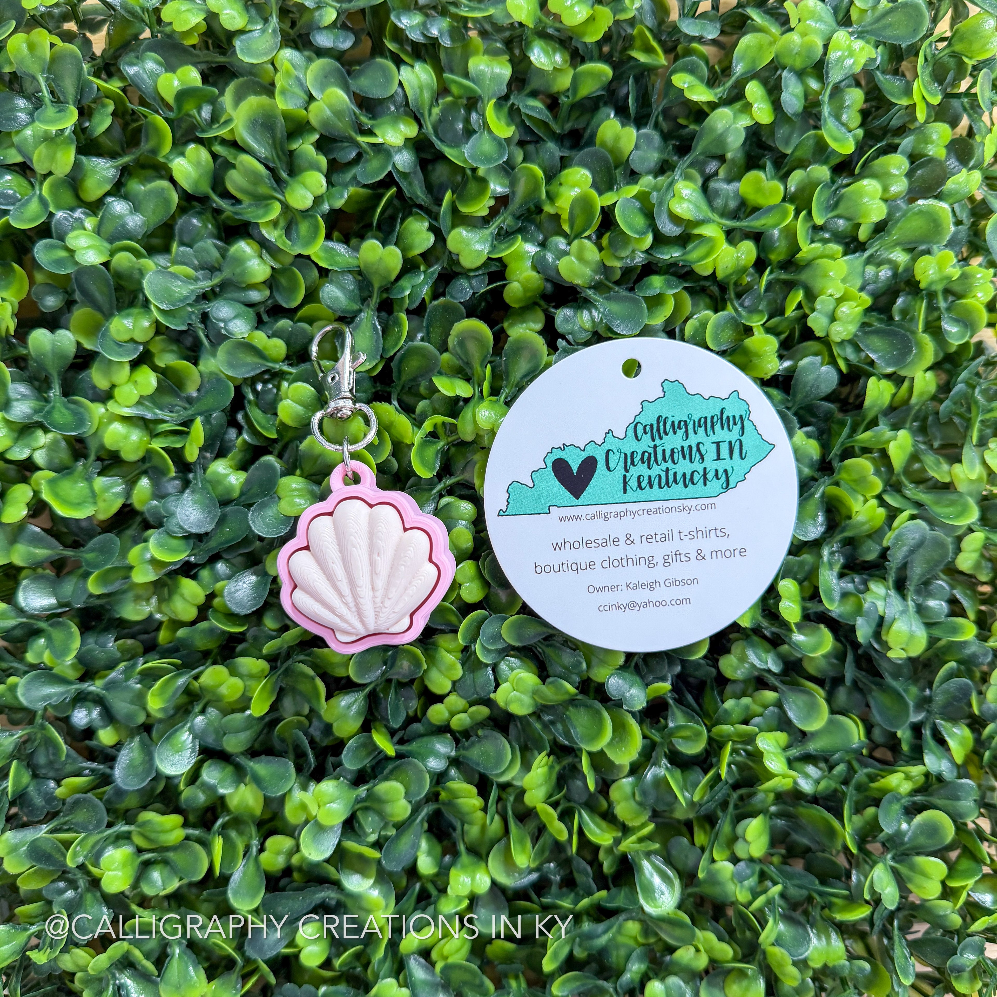Pink shell-shaped fidget clicker keychain with a tag on a green leafy background