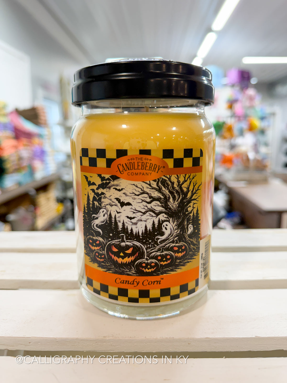 A jar candle labeled 'Candy Corn' by Candleberry Company, featuring a black lid and a Halloween-themed label with pumpkins, a ghost, and a spooky tree, set on a white wooden shelf in a store.