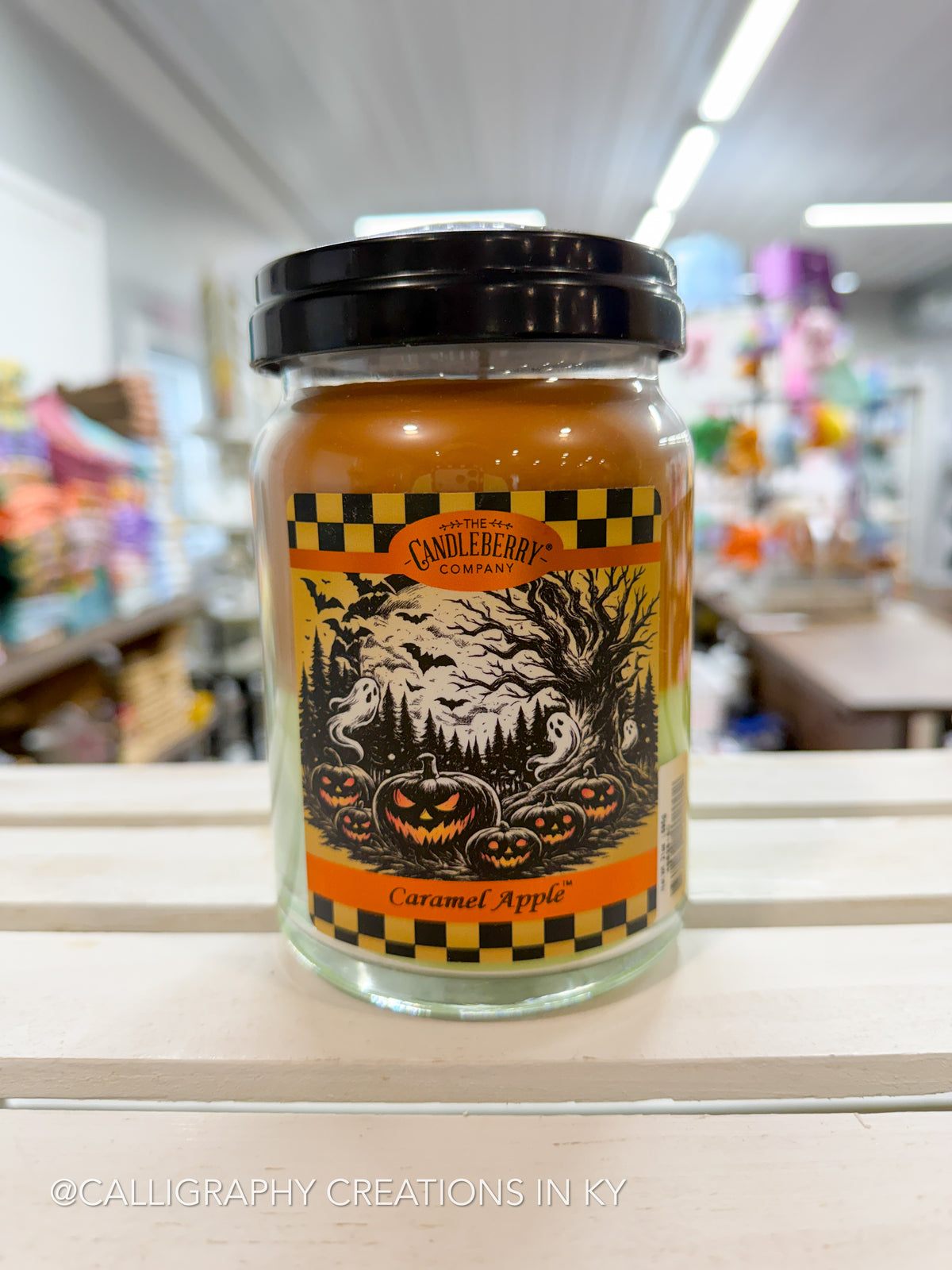 A CandleHero 'Caramel Apple' scented candle in a clear glass jar with a black lid, set on a white wooden surface with a blurred store background.