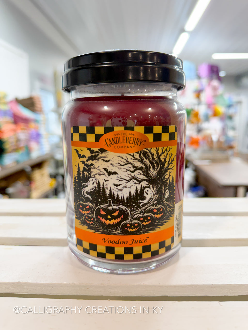 A jar candle labeled 'Wooden Juice' by Candleberry Company, featuring a black lid and a Halloween-themed label with pumpkins, ghosts, and bats, set on a wooden shelf in a store.