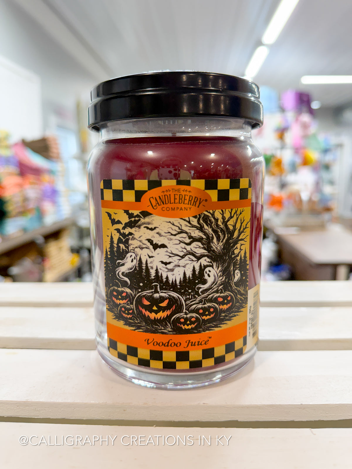 A jar candle labeled 'Wooden Juice' by Candleberry Company, featuring a black lid and a Halloween-themed label with pumpkins, ghosts, and bats, set on a wooden shelf in a store.