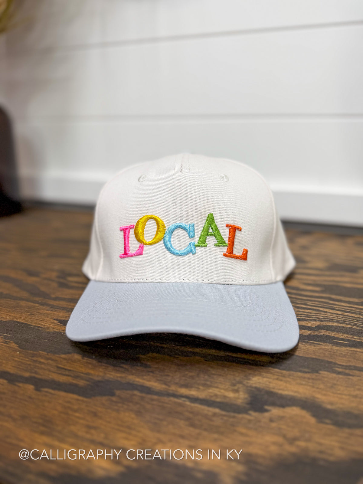A white baseball cap with a light gray brim, featuring the word 'LOCAL' embroidered in colorful letters across the front. The cap is placed on a dark wood surface with a white paneled wall in the background.