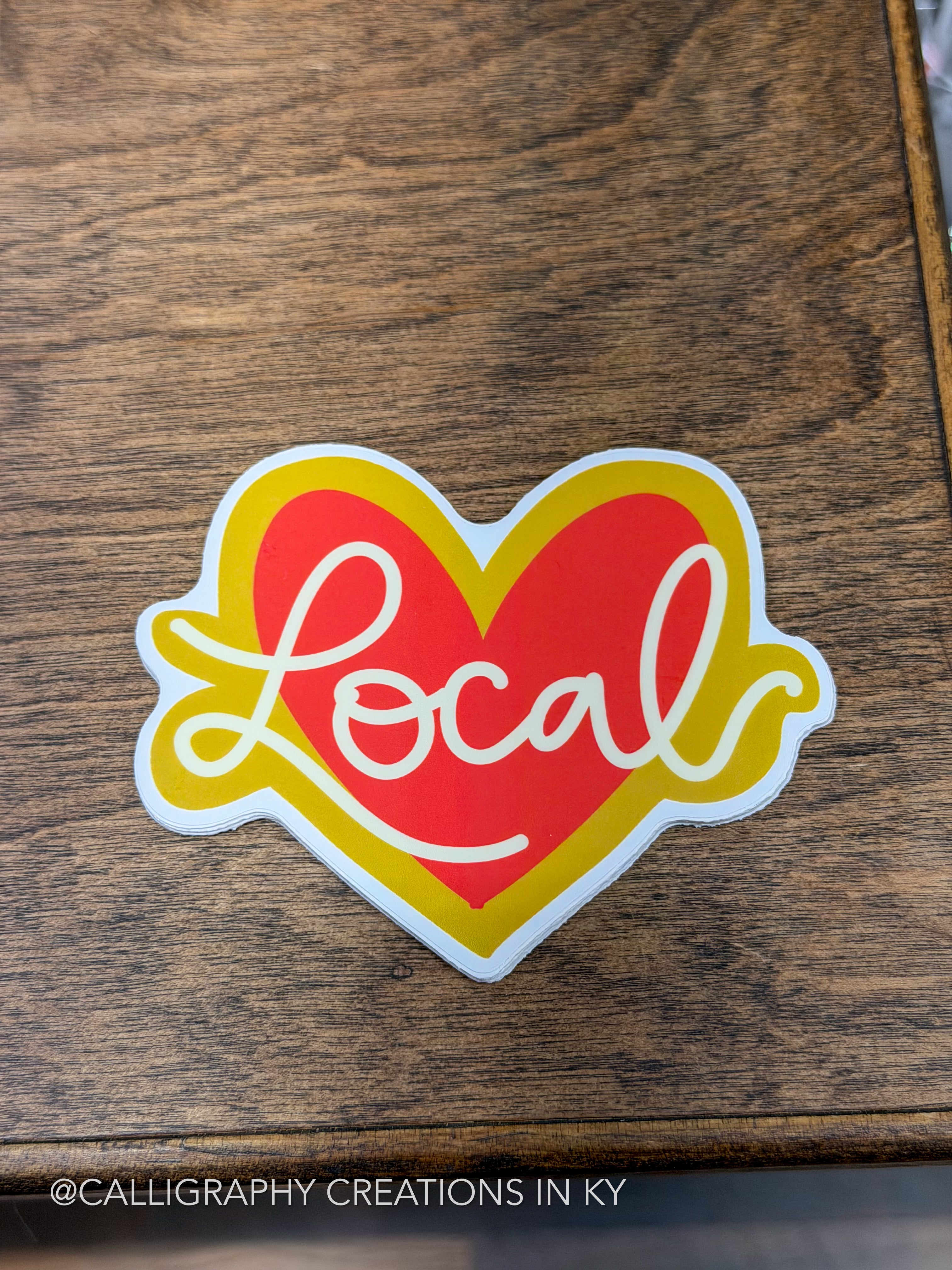 A die-cut sticker featuring a red heart with a yellow outline and the word 'Local' in white cursive script, placed on a dark wood surface.
