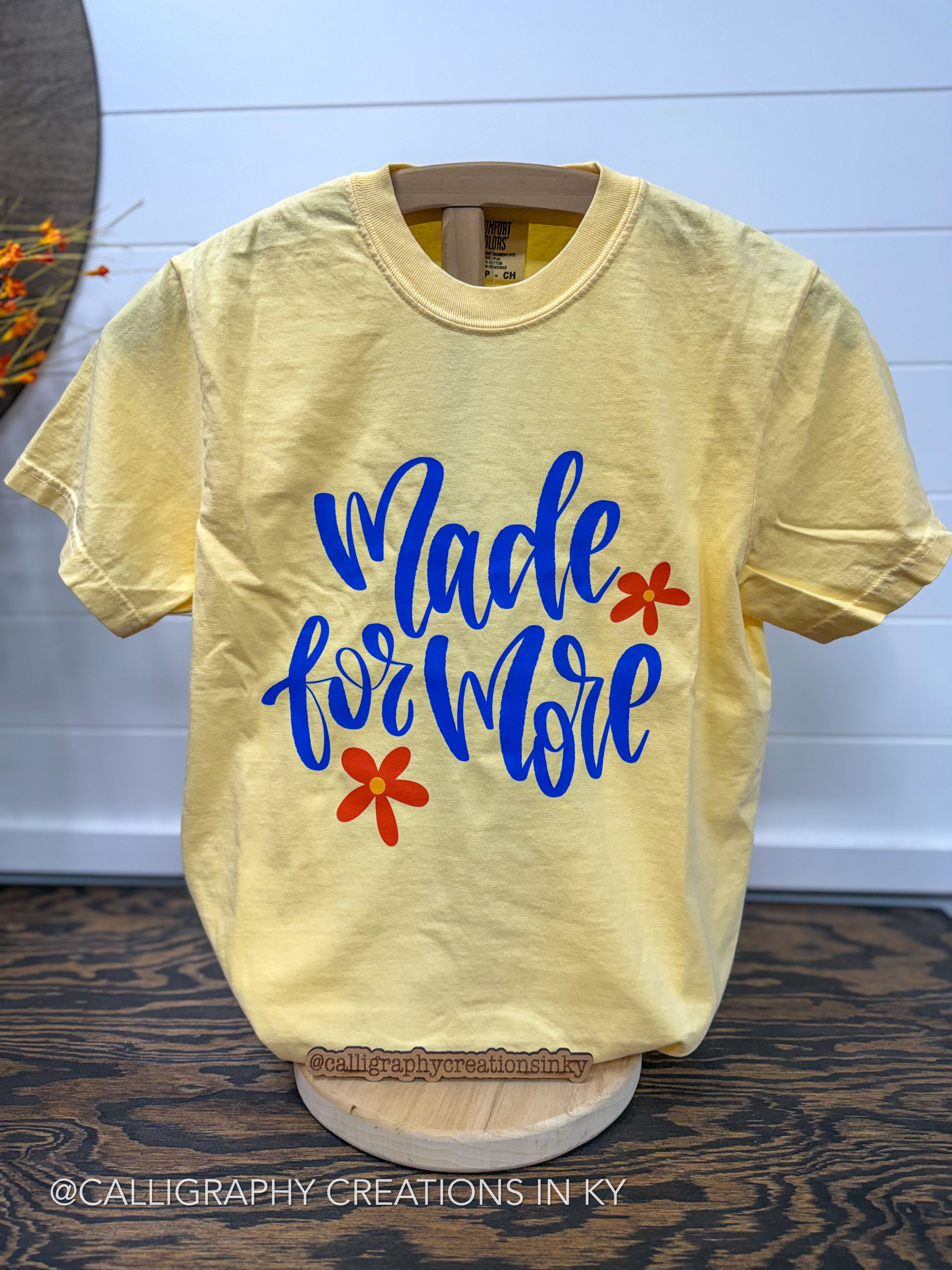 A pale yellow graphic T-shirt displayed on a wooden hanger and stand, featuring the phrase 'Made for More' in blue script with two small red flower graphics. The shirt is photographed indoors against a white paneled wall above a dark wood surface.