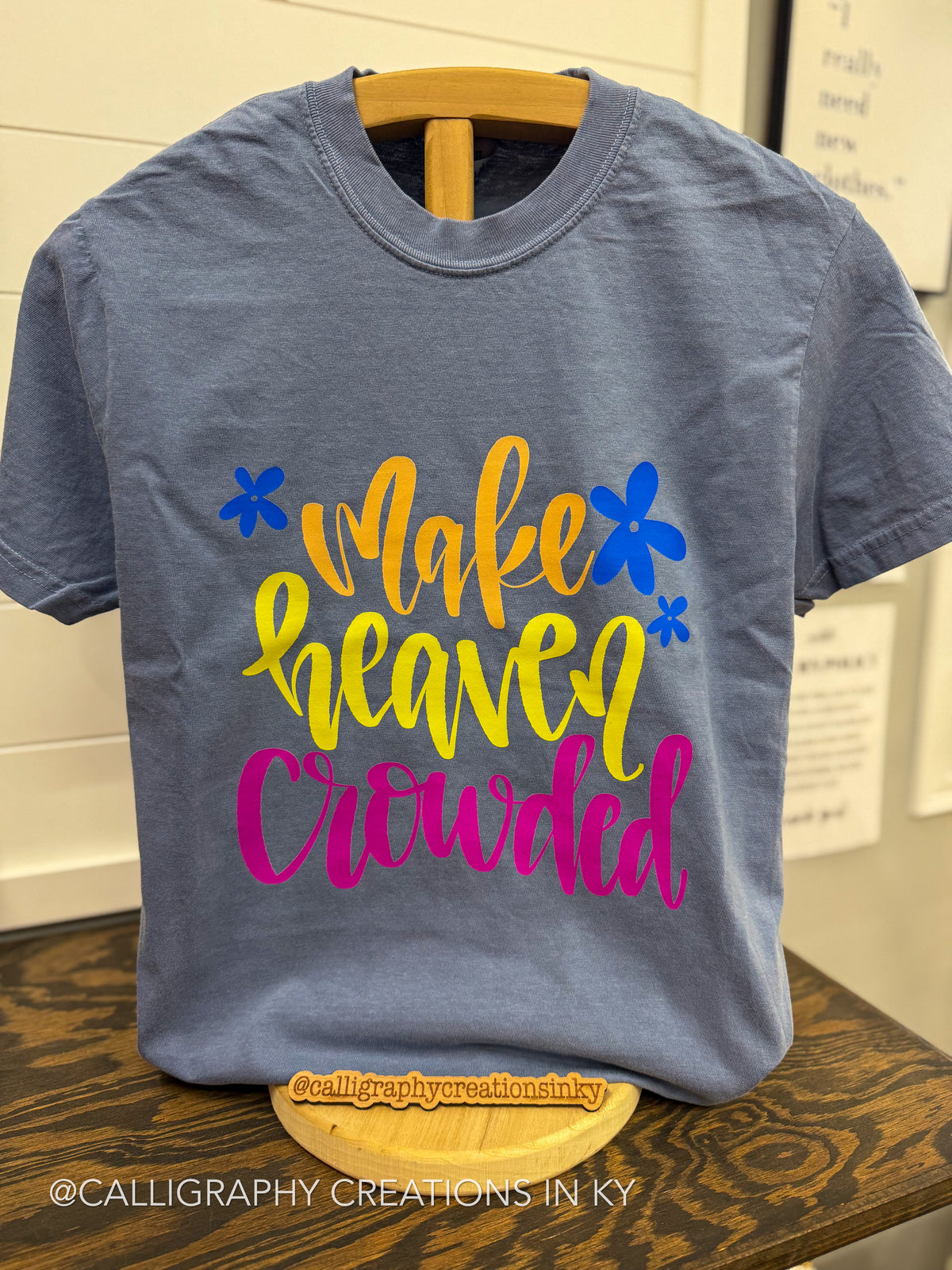 A slate blue graphic t-shirt displayed on a wooden hanger on a rustic wooden table. The shirt features the phrase 'Make Heaven Crowded' in stylized yellow, orange, and pink lettering, accented with blue flower graphics.